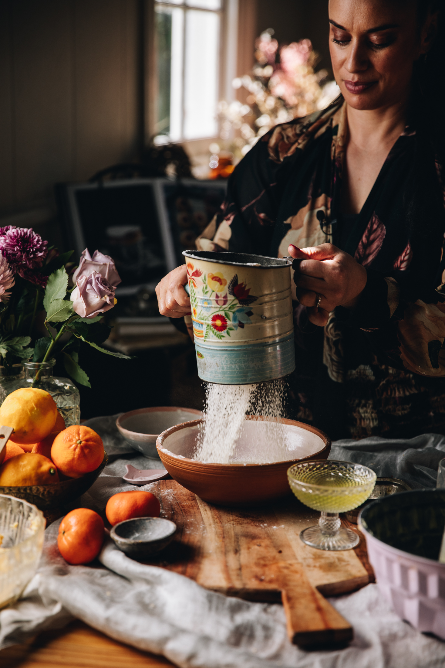 Naomi Toilalo is holding an old blue vintage sifter and is sifting flour in to a ceramic bowl. On the table next to her is the flowers and citrus. 