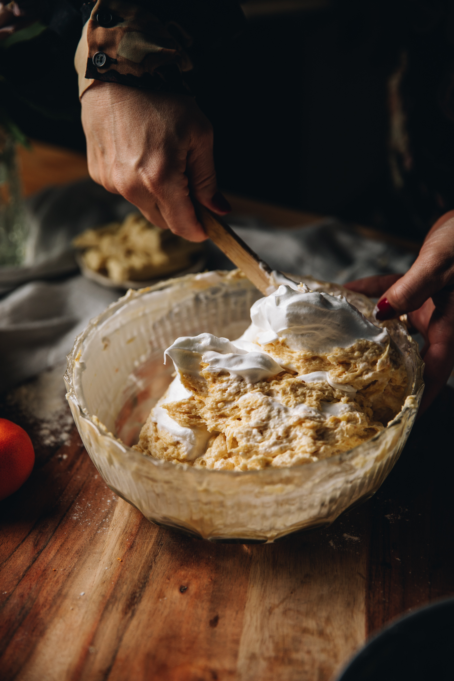 A glass vintage bowl sits on a wooden table. It has a cake mixture in it and meringue is being folded in to it. Streaks of meringue can be seen in the mixture. 