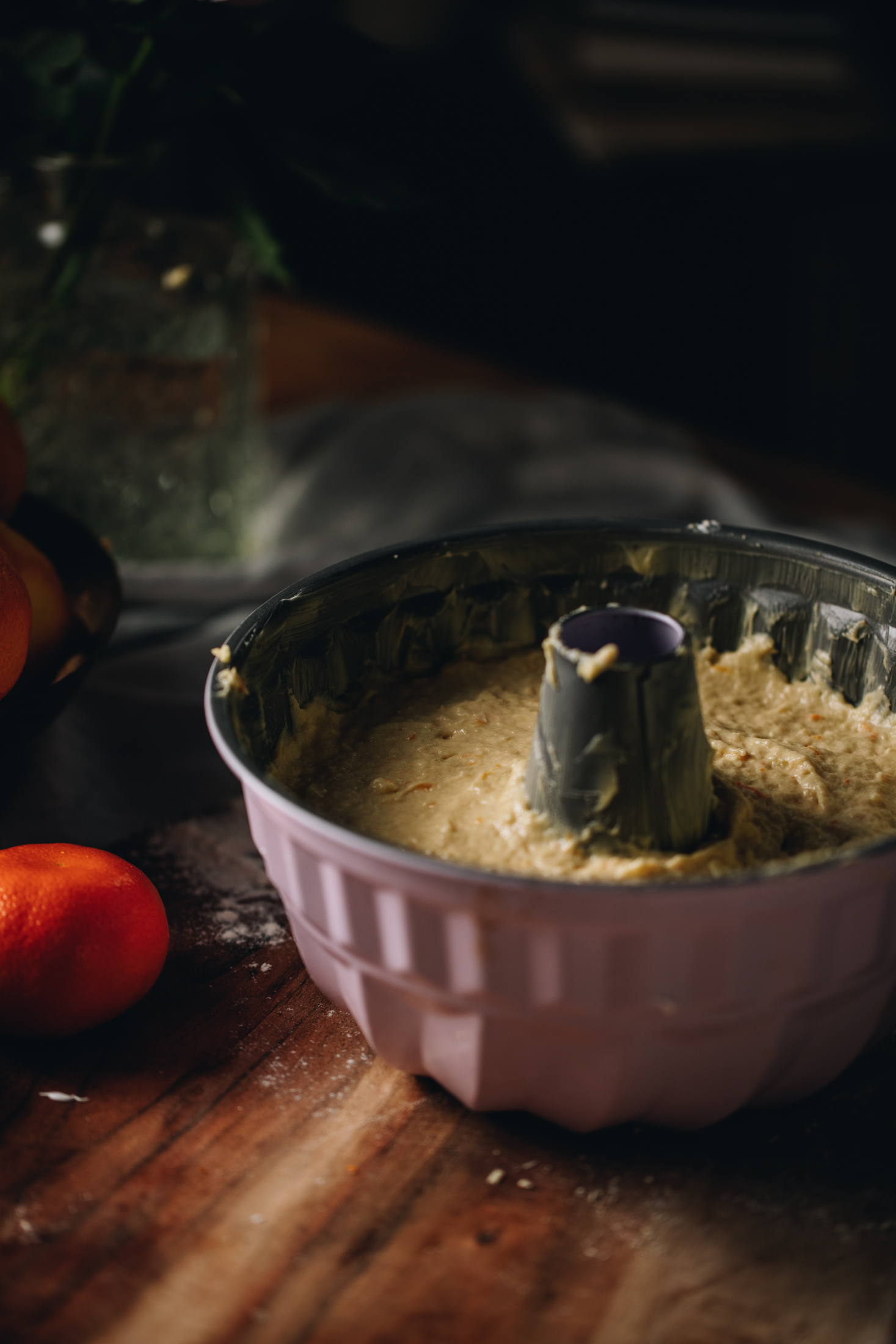 A rose pink bundt cake tin sits on a wooden. It has been filled with cake batter. There is a mandarin next to it. 