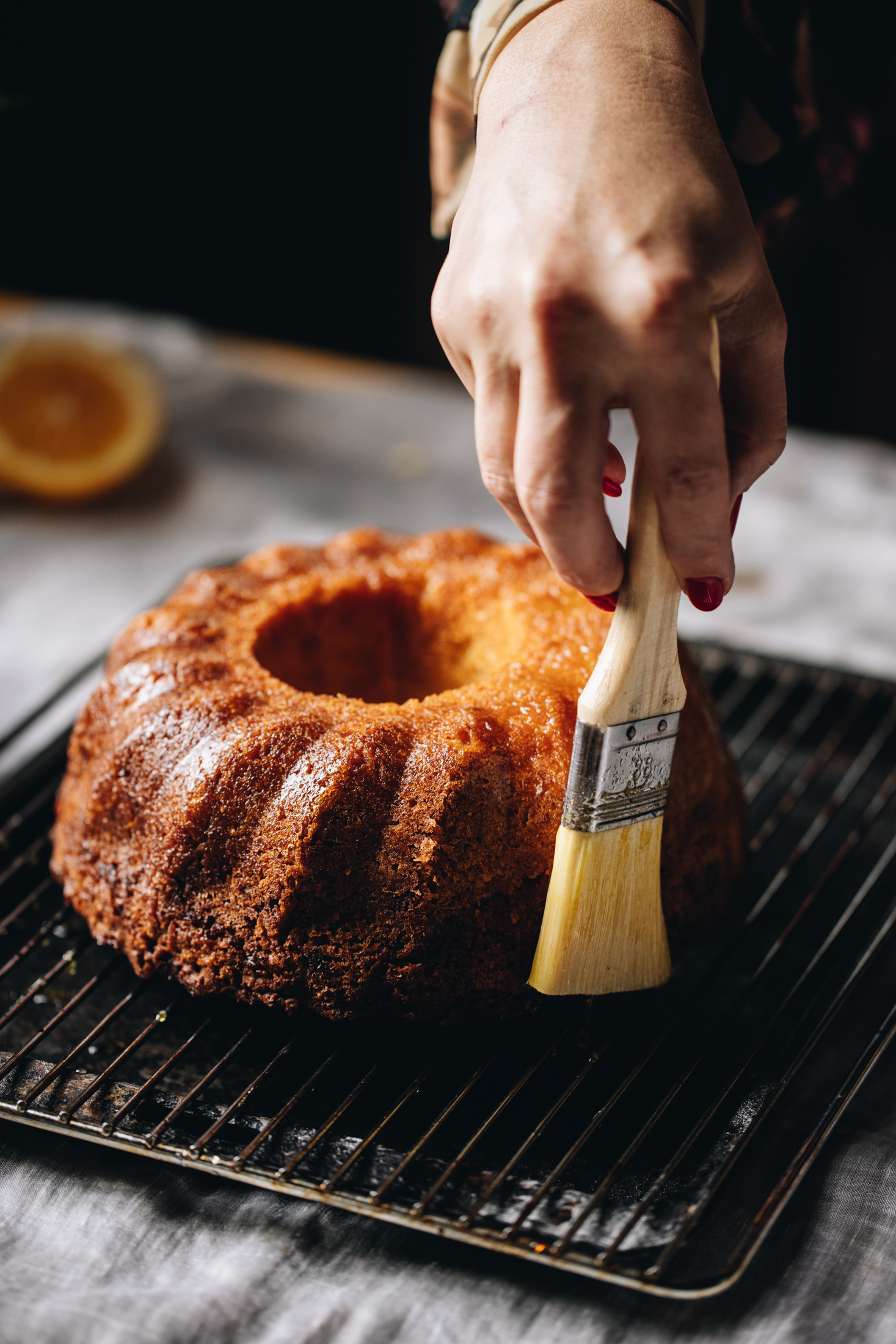 On a soft grey tablecloth sits a black tray that has a cooling rack on top of it. On top of the cooling rack is a freshly baked Lemon Syrup Bundt Cake that is being basted with lemon syrup with a pastry brush. 