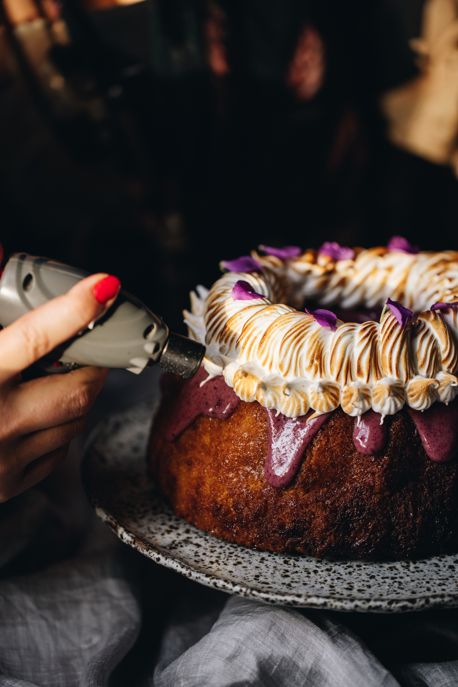Sitting on a ceramic cake stand is lemon syrup bundt cake. It is decorated with a boysenberry ganache dripping down the sides and is topped with piped meringue that is being torched in the shot. 
