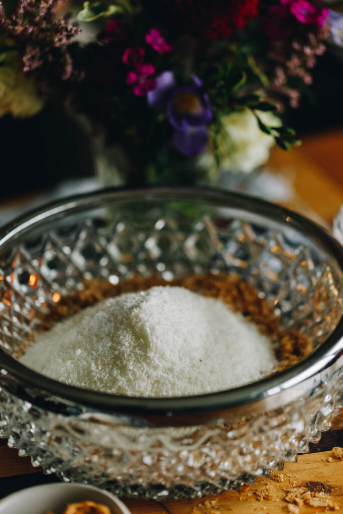 A glass bowl with a silver trim is sitting on a wooden board with coconut in a pile inside it. Flowers are in the background. 
