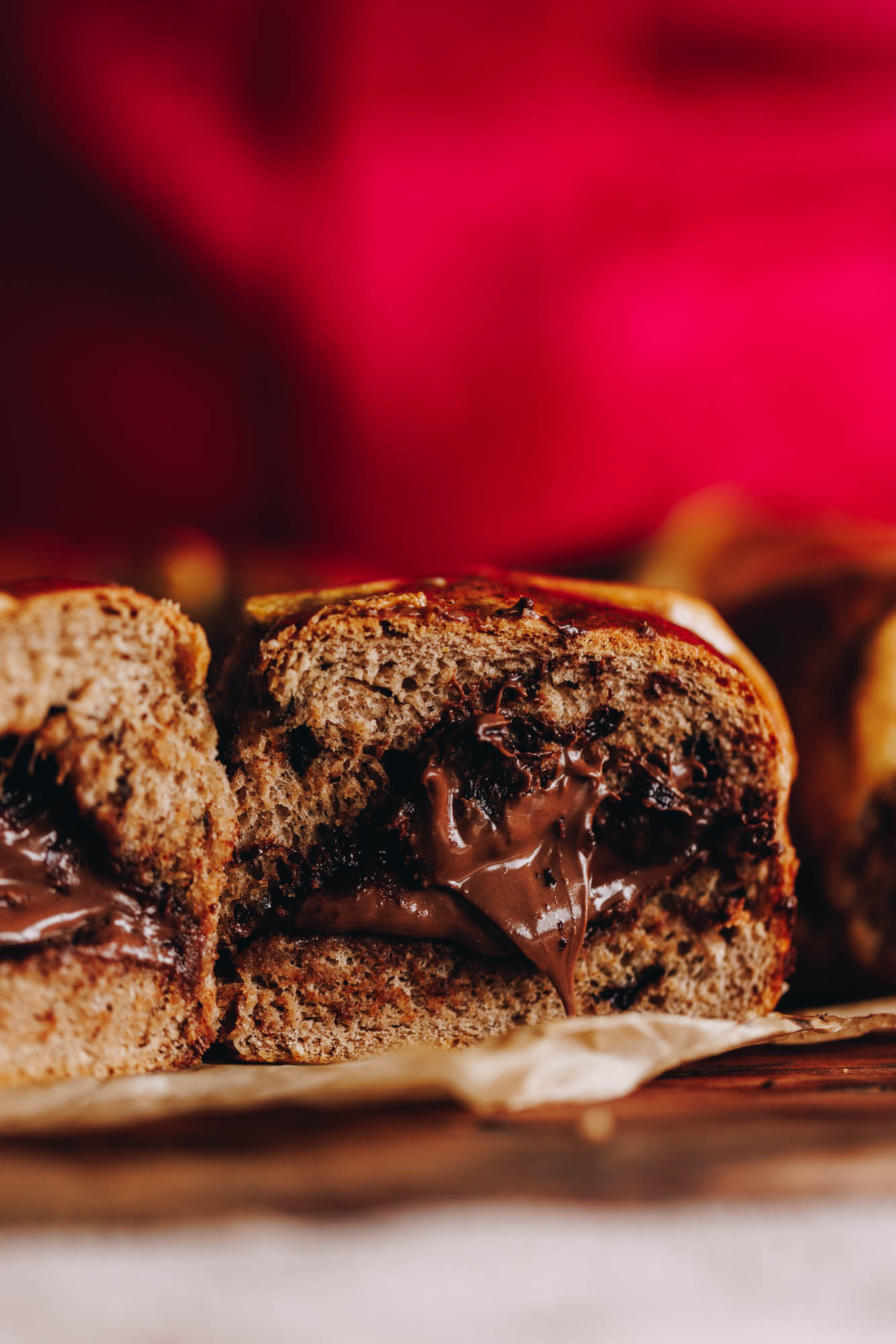 A close up shows two freshly baked Nutella hot cross buns side by side. They have been cut open and reveal the gooey Nutella coming out of the centre. It has a bright pink backdrop. 