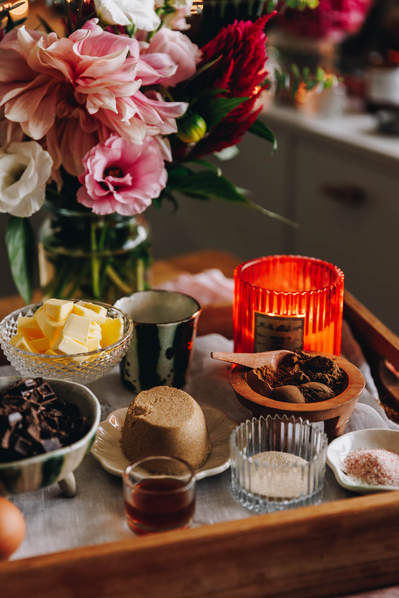 A wooden tray sits on a table. In the tray is a soft cream fabric. On the fabric is different vintage vessels holding ingredients. There is salt, yeast, vanilla, brown sugar, spices, chocolate and butter in view. An orange candle is also burning behind th spices. A glass vase of beautiful pink and peach flowers is behind the wooden tray. 