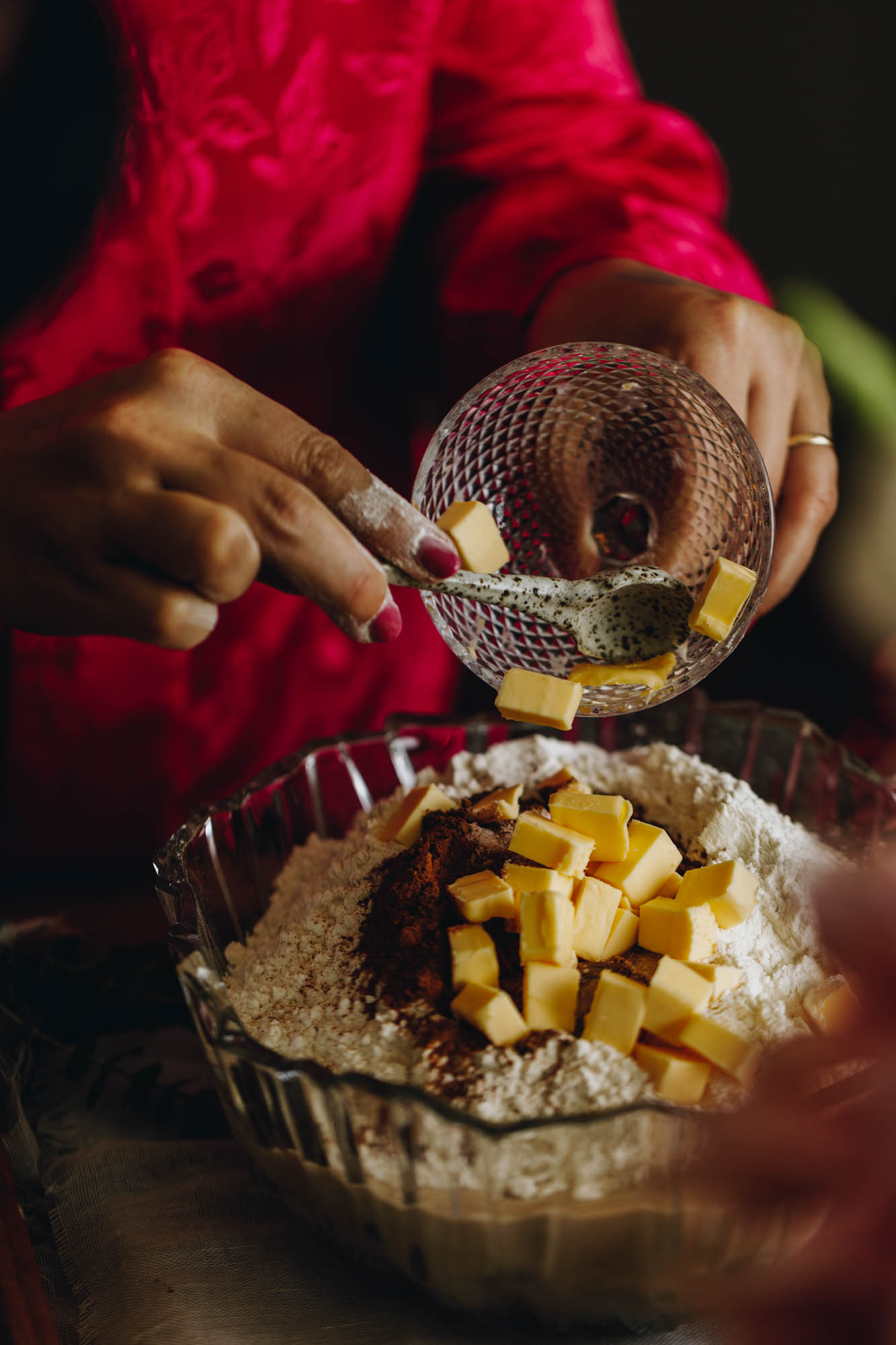 A vintage glass bowl sits on a wooden board. In the bowl is a flour mixture with spices in it. Naomi is adding cubed butter on top out of a small glass. 
