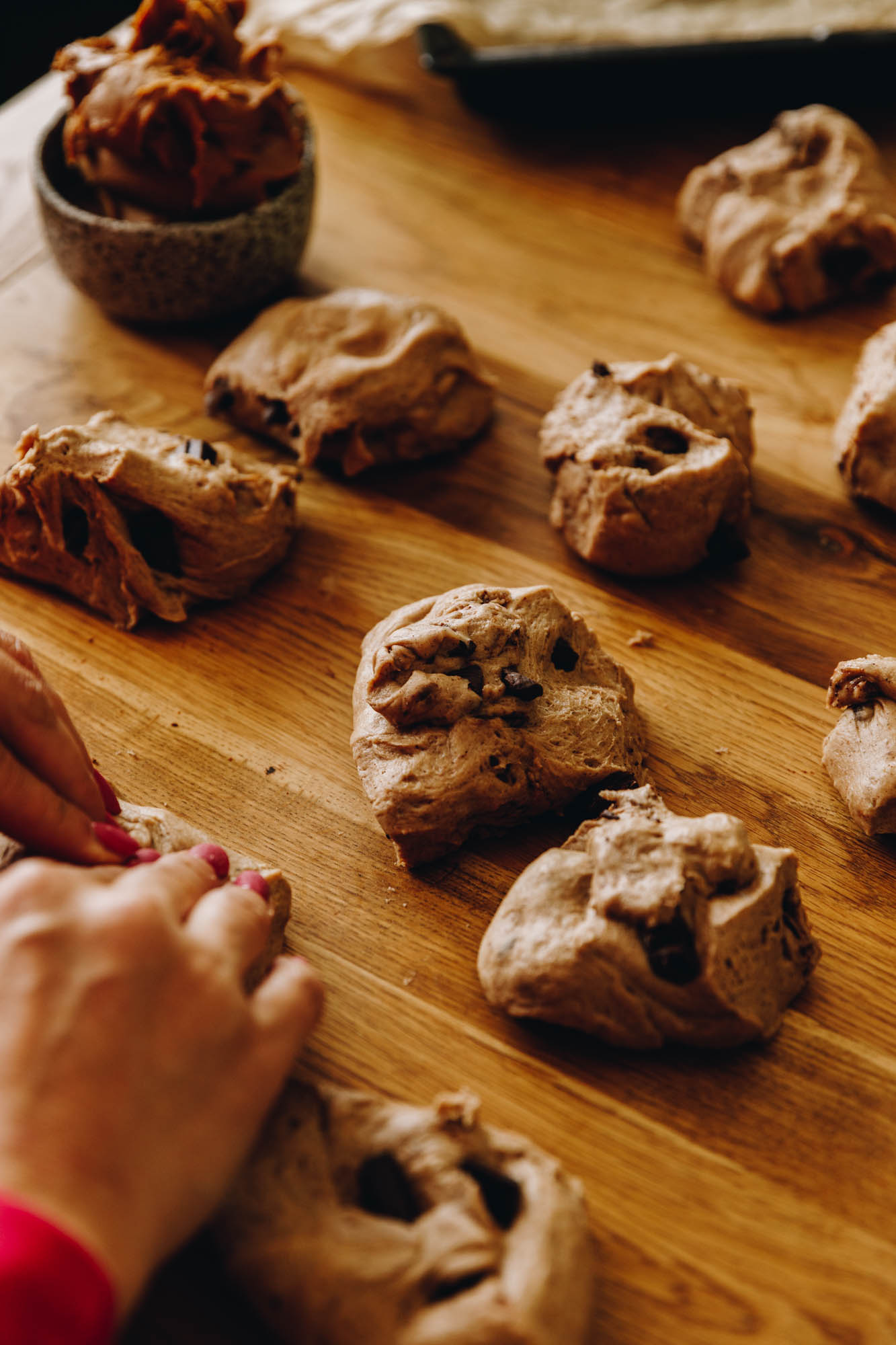 On a wooden table is blobs of spiced dough with chocolate chunks in them. Naomi is pressing one of the dough with her hands. 