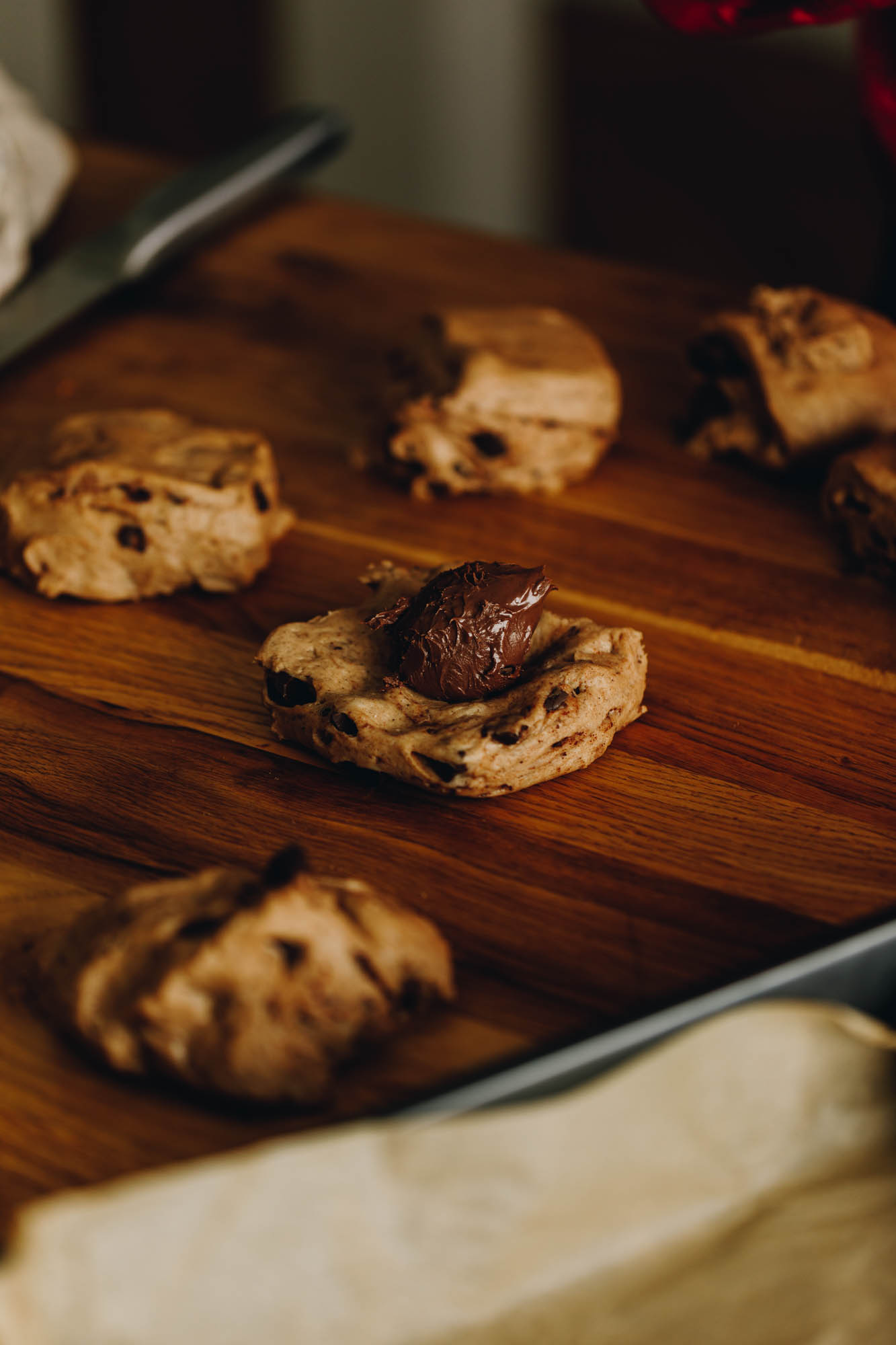 On a wooden table is five blobs of heavily spiced dough studded with dark chocolate chunks. One of the dough pieces has been flattened out and a big blob of Nutella sits in the centre. A tray with brown baking paper is at the front of the shot. A large silver knife is in the background. 