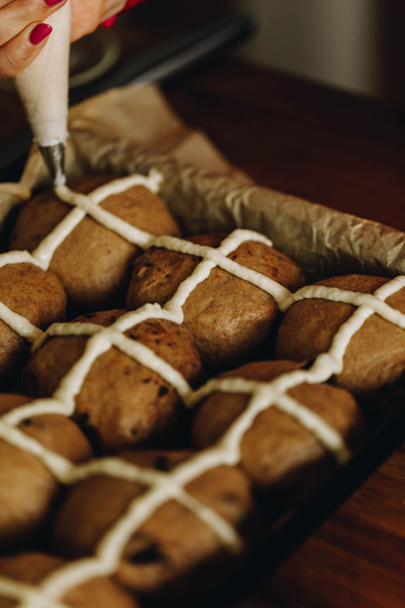 A black tray lined with brown baking paper sits on a wooden table. In the tray is unbaked hot cross buns and the crosses are being piped on with a piping bag. 