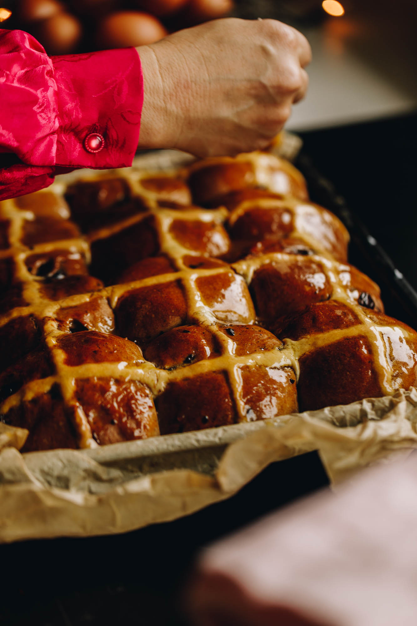Baked Nutella hot cross buns are in a black tray, lined with brown baking paper. The buns are being glazed with sticky syrup which makes the buns glossy. 