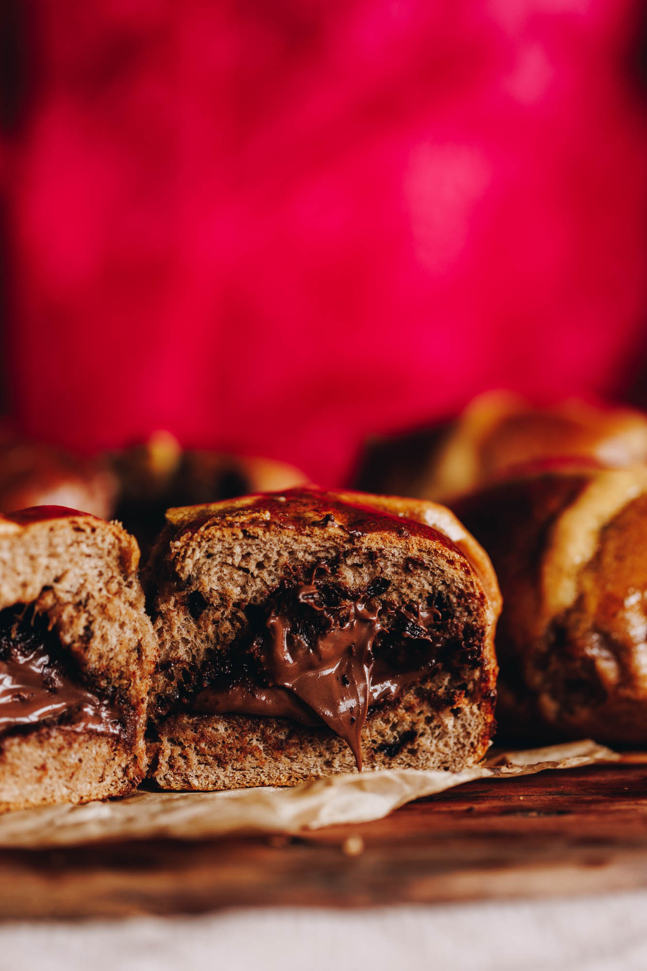A close up shows two freshly baked Nutella hot cross buns side by side. They have been cut open and reveal the gooey Nutella coming out of the centre. It has a bright pink backdrop.