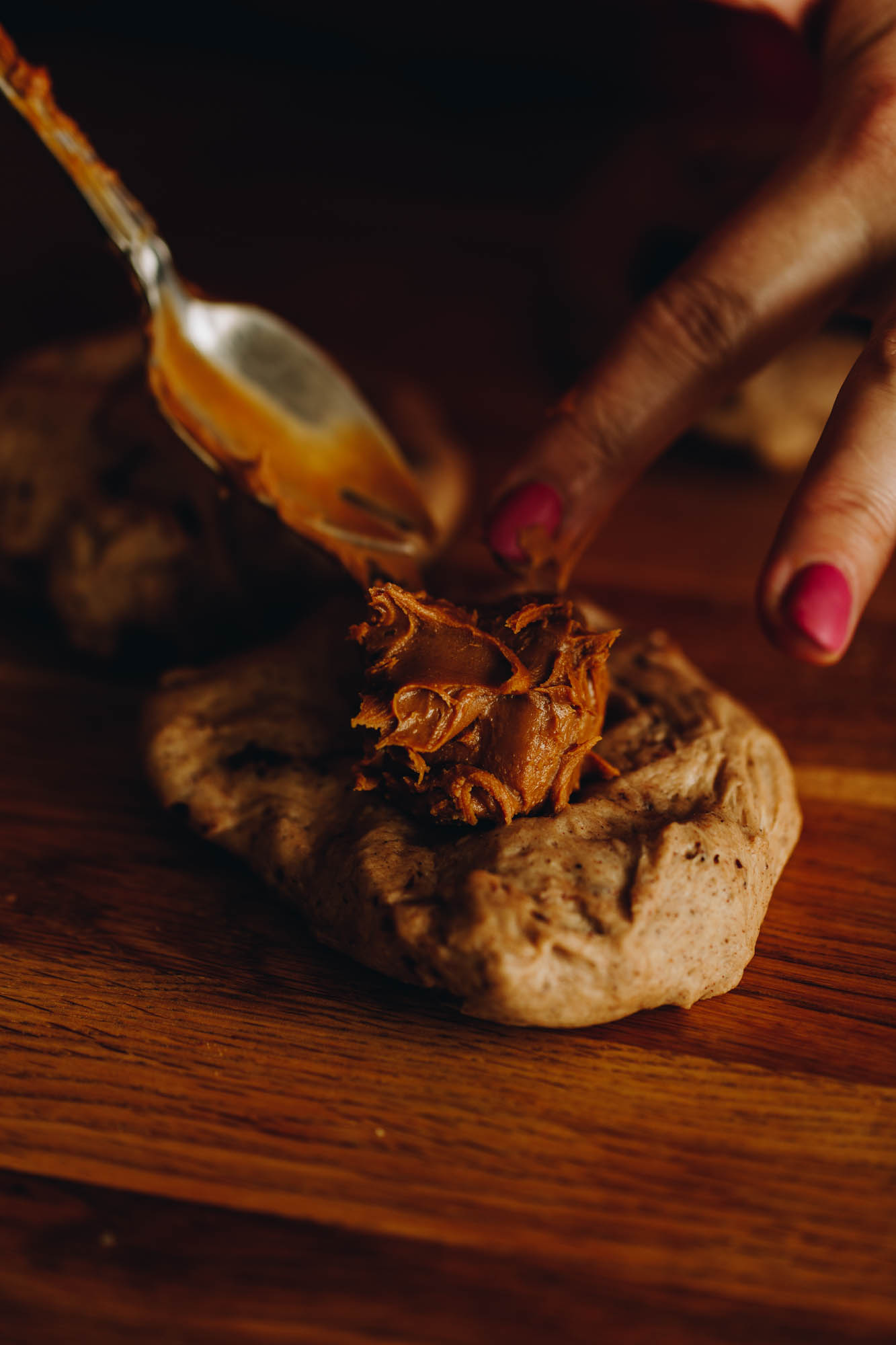 A close up shots shows a piece of spiced dough on a wooden table and a teaspoon adding a large blob of biscoff in to the centre. 