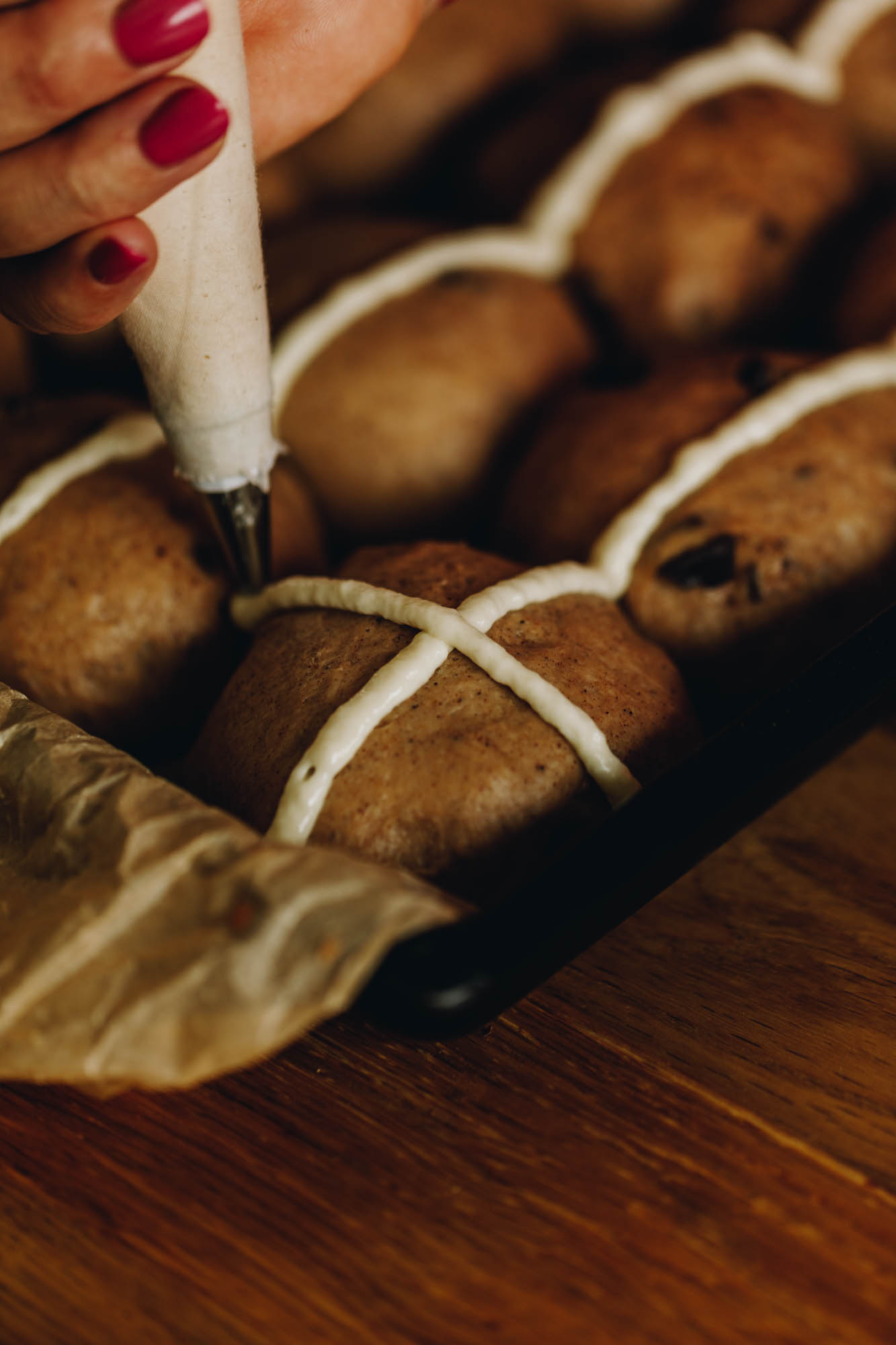 A black tray lined with brown baking paper sits on a wooden table. In the tray is unbaked hot cross buns and the crosses are being piped on with a piping bag.