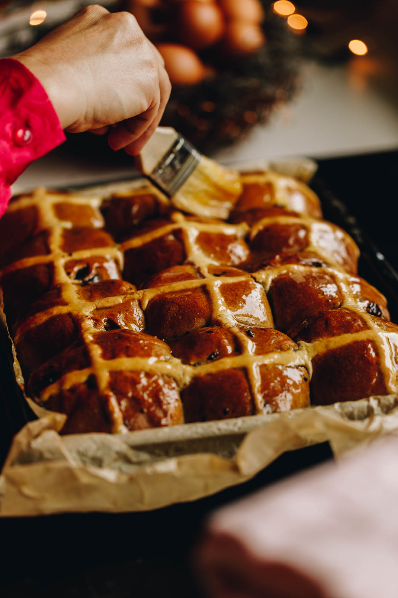 Baked Biscoff hot cross buns are in a black tray, lined with brown baking paper. The buns are being glazed with sticky syrup which makes the buns glossy.
