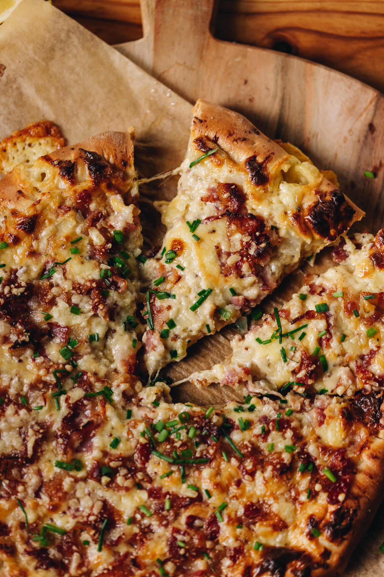 A baked cheese and onion pizza sits on a brown baking paper on a round wooden board, that sits on a wooden table. One of the slices of pizza has been cut and cheese is pulling from the sides of the slice. There is bacon, chives and melty cheese all over the pizza. 