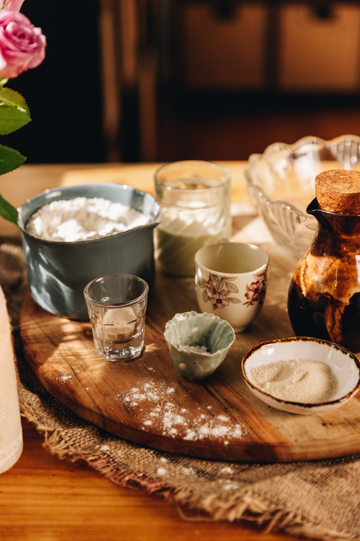 A wooden board sits on a natural fabric, on a wooden table. On the round board is ingredients that are in vintage vessels. There is yeast, sugar, flour, milk, a ceramic jug and a glass bowl in view. Pink flowers are in the background. 
