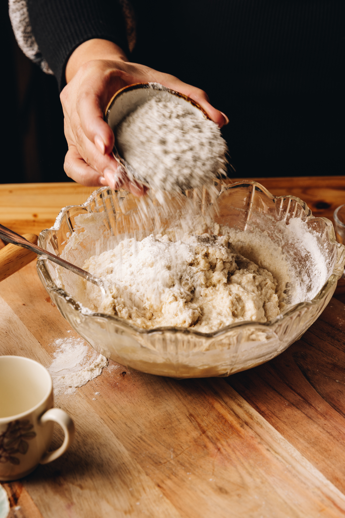 A vintage glass bowl sits on a wooden table. In the bowl is dough ingredients and Naomi is adding flour on top of it from a small ceramic bowl. A small cup is next to the bowl.