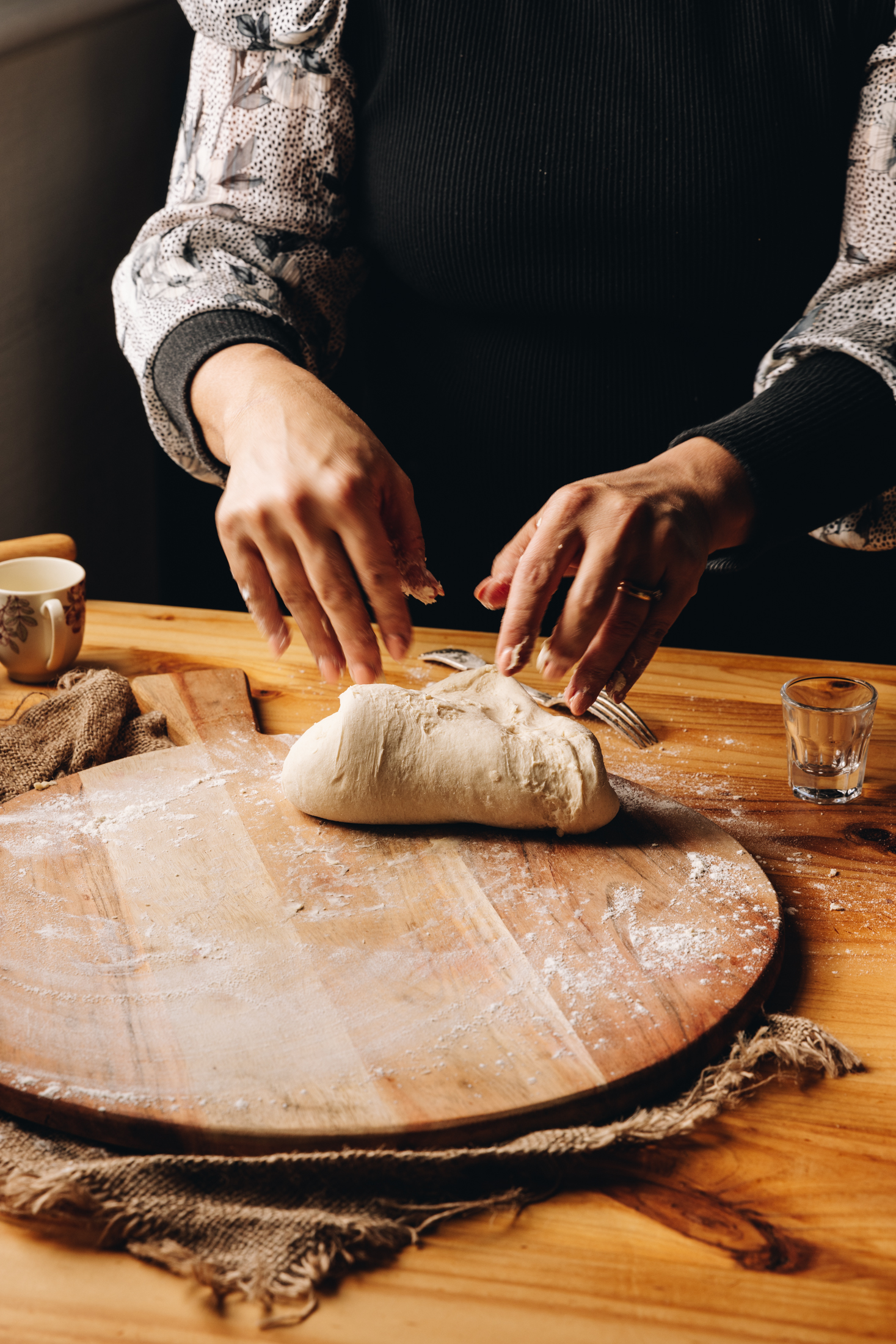 A wooden board sits on a natural fabric, on a wooden table. On the round board is flour and a dough that Naomi is kneading with her hands.