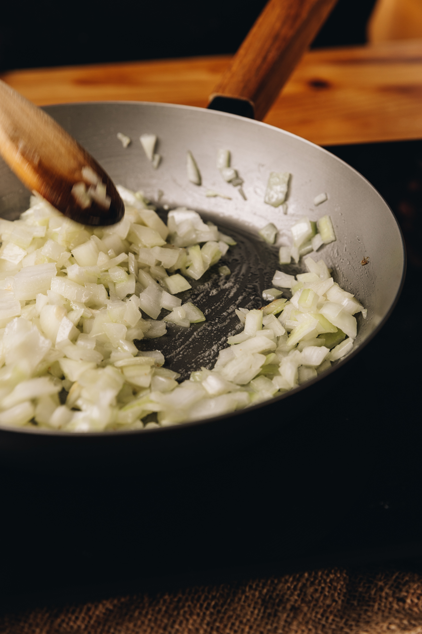 A silver cast iron pan sits on a black portable stove. In the pan is diced onions that are being stirred with a wooden spoon. 