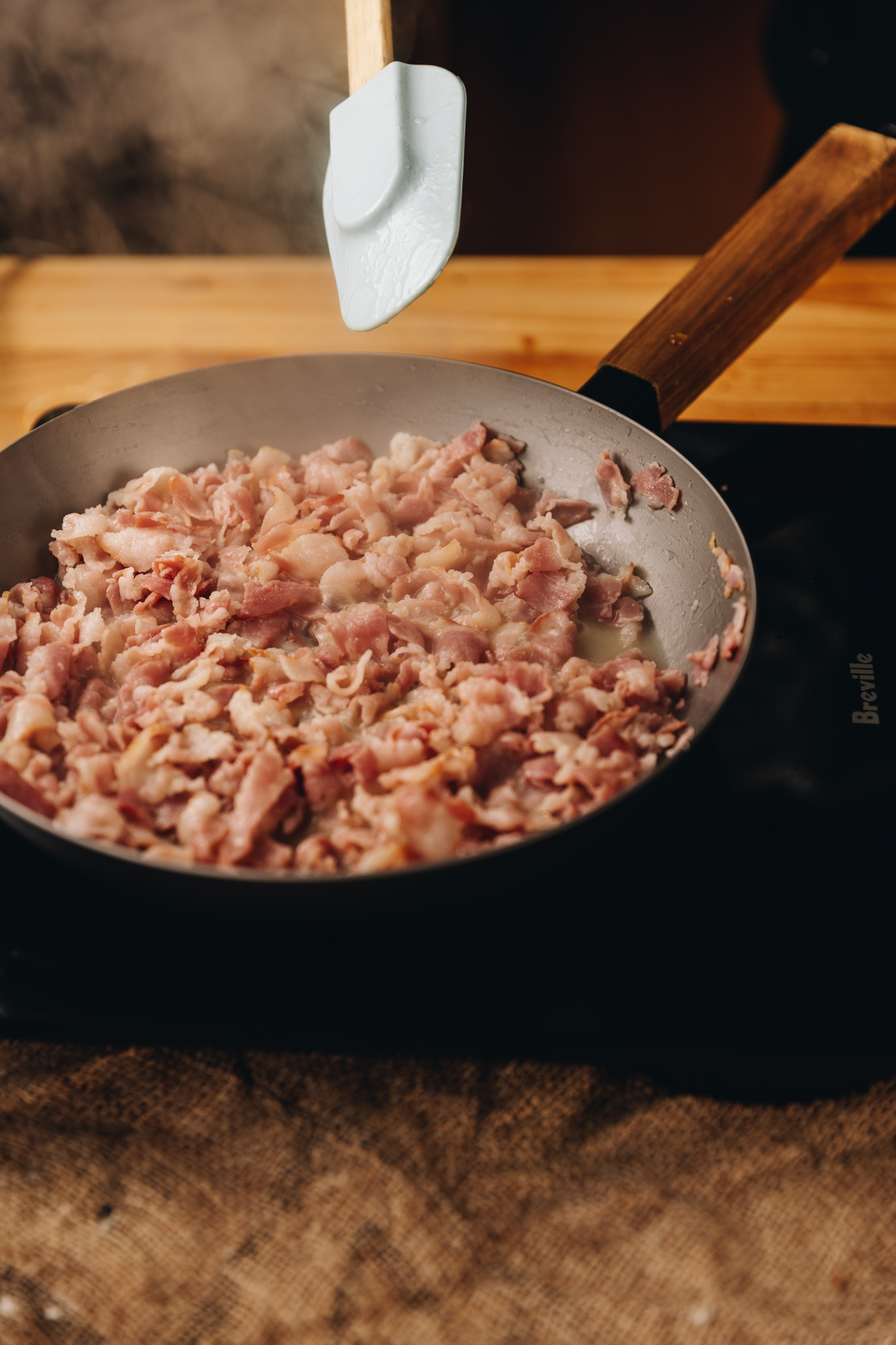 A silver cast iron pan sits on a black portable stove. In the pan is bacon pieces that are steamy. A blue spatula with a wood handle is coming in to frame, to stir it. 