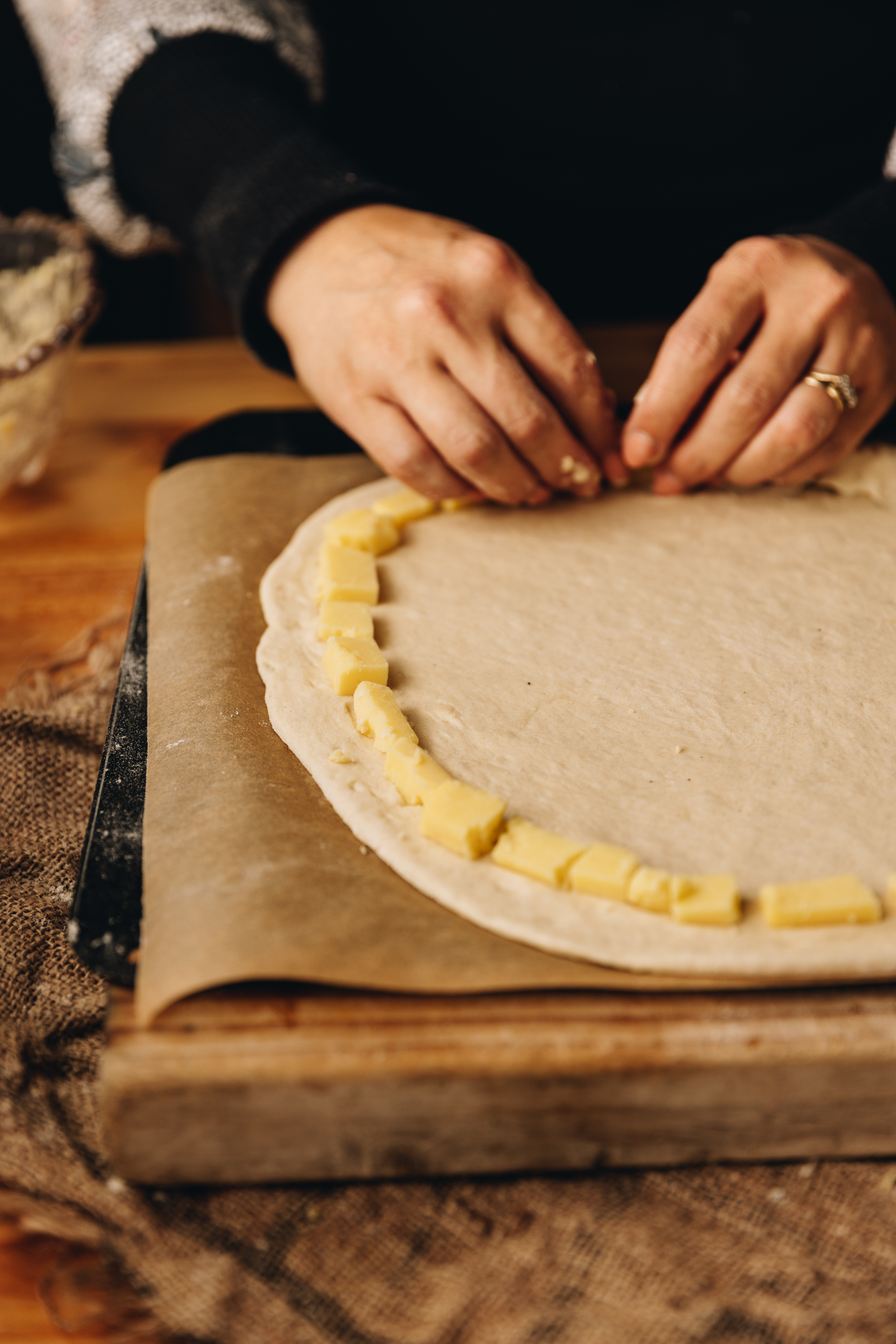 A wooden board is on a wooden table. It is lined with brown baking paper and has a rolled out pizza dough on it. Around the edge are small pieces of cheese that line the crust. 