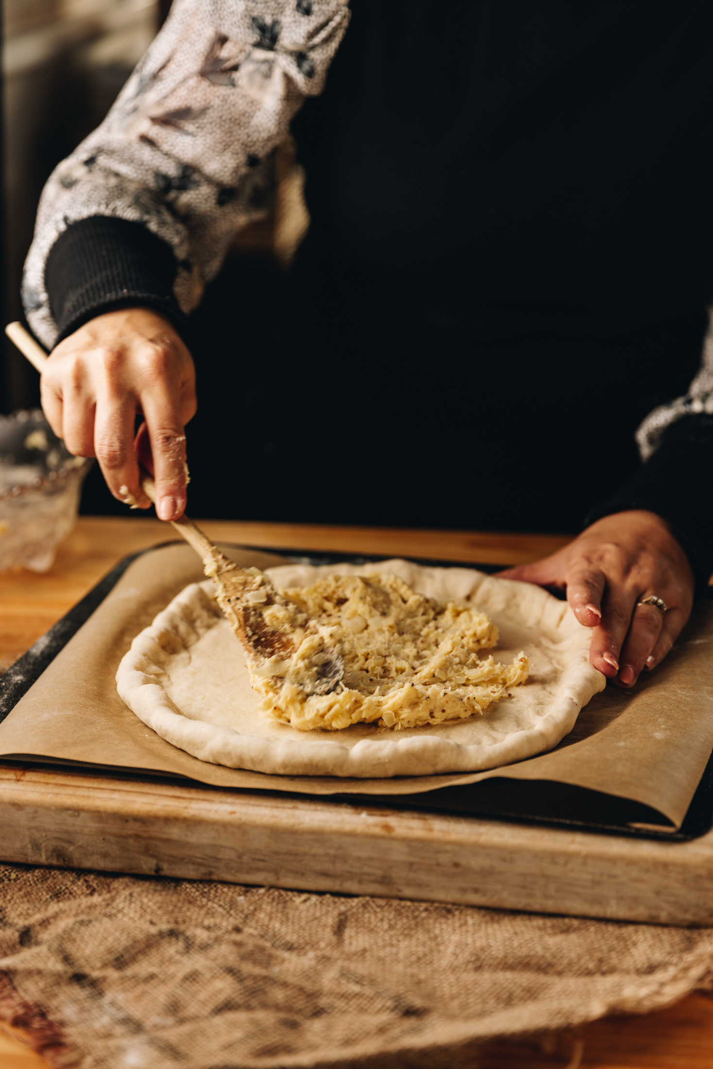A wooden board is on a wooden table. It is lined with brown baking paper and has a rolled out pizza dough on it which has been stuffed with cheese. Naomi is spreading the pizza with a thick cheese sauce, using a wooden spoon. 