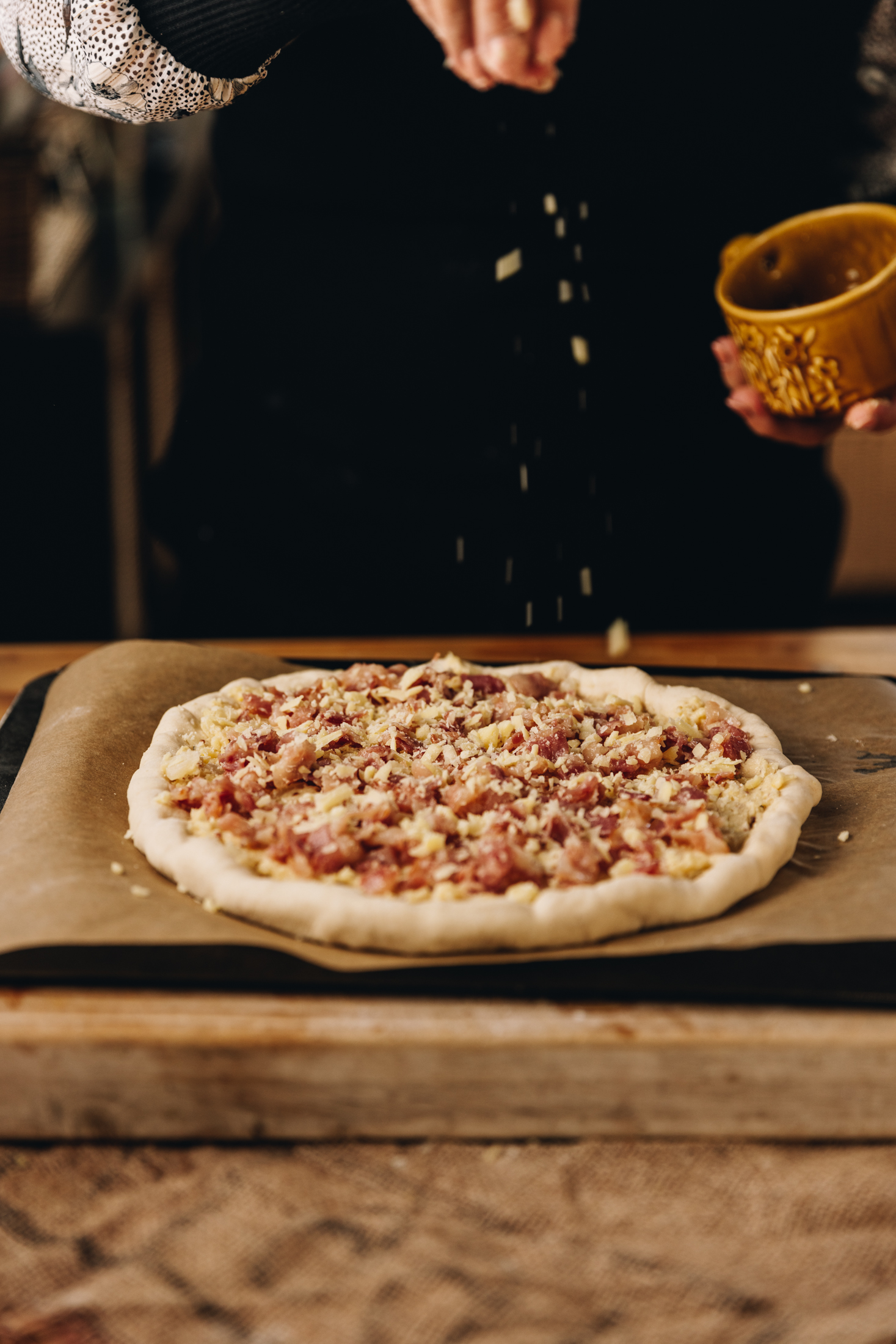 A wooden board is on a wooden table. It is lined with brown baking paper and has a rolled out pizza dough on it which has been stuffed with cheese. There is bacon all over it. Naomi is now sprinkling grated cheese on to it. 