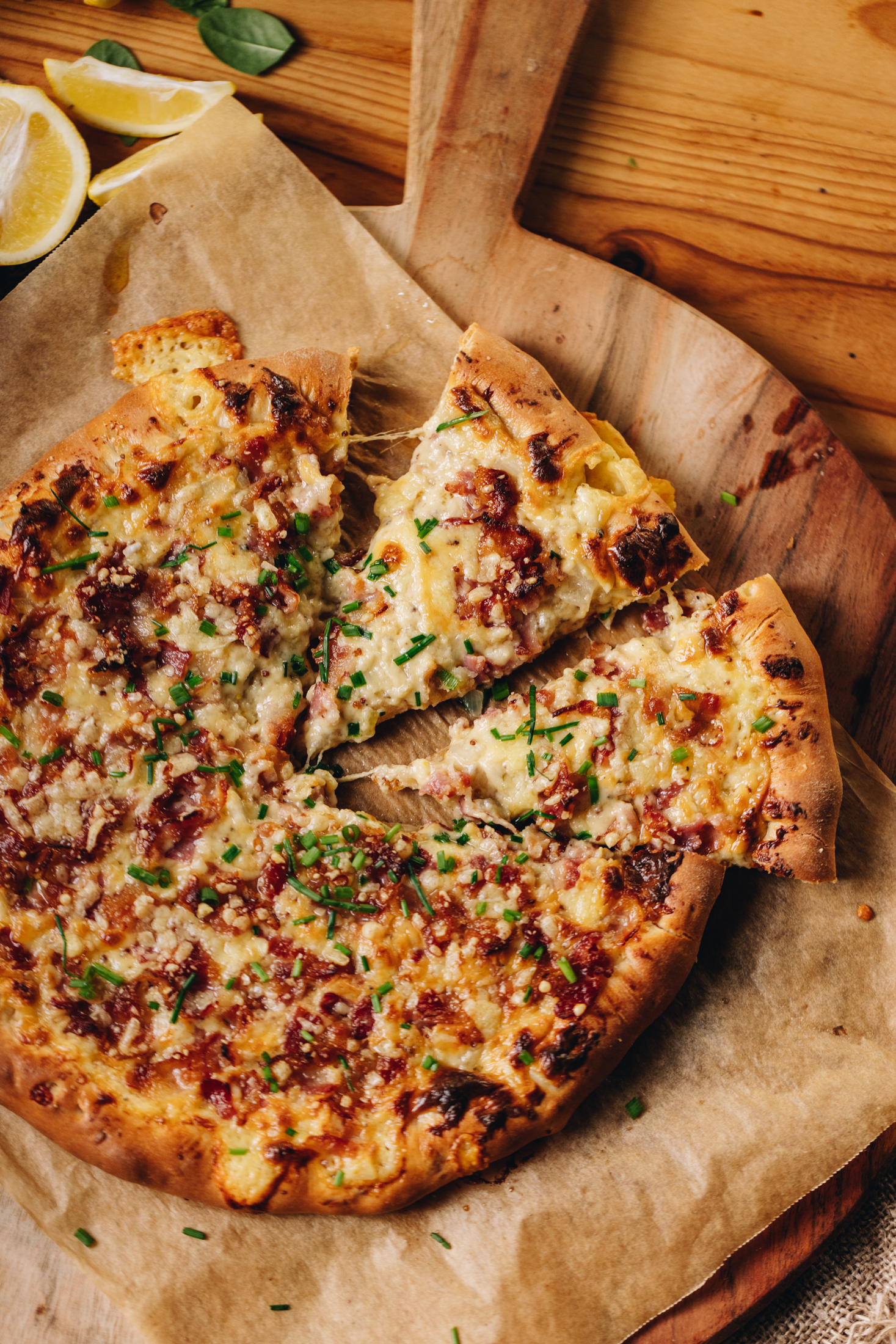 A baked cheese and onion pizza sits on a brown baking paper on a round wooden board, that sits on a wooden table. Two of the slices of pizza has been cut and cheese is pulling from the sides of the slices. There is bacon, chives and melty cheese all over the pizza.