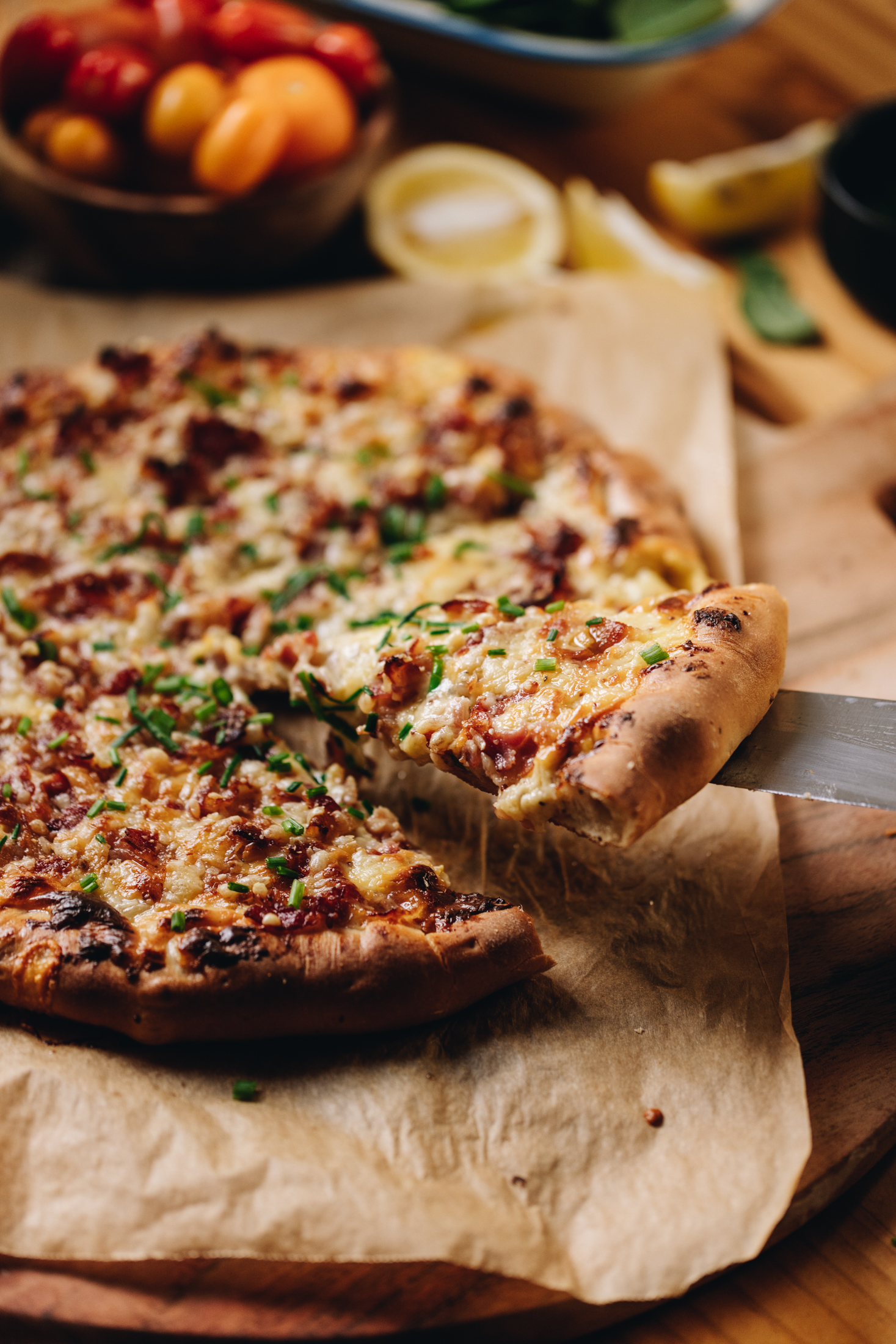 A freshly baked cheese and onion pizza is on a tray lined with brown baking paper. A knife is lifting off a slice and cheese is pulling off the piece. There is lemons and tomatoes in the background. 
