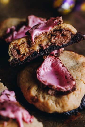 An easter egg cookie is on a wooden board, another cookie has been cut in half to reveal the gooey inside.