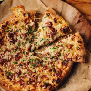 A top shot shows A whole cheese and onion pizza on a round wooden board, two pieces are cut and pulled out revealing the gooey cheese topping.