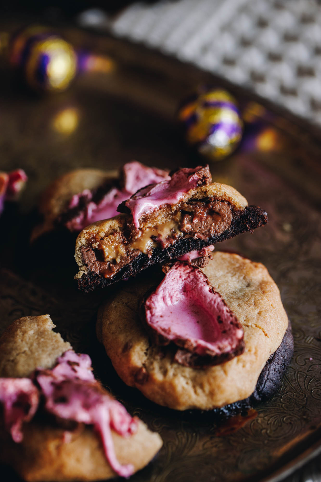 Freshly baked Chocolate easter egg cookie sit on a gold tray. One is whole and another one has been cut in half to reveal the gooey chocolate centre. The cookies are two tone, the top layer is vanilla and the bottom layer is chocolate. On top of the cookies is melted pink marshmallow. Mini easter eggs and a weaved mat are in the background.