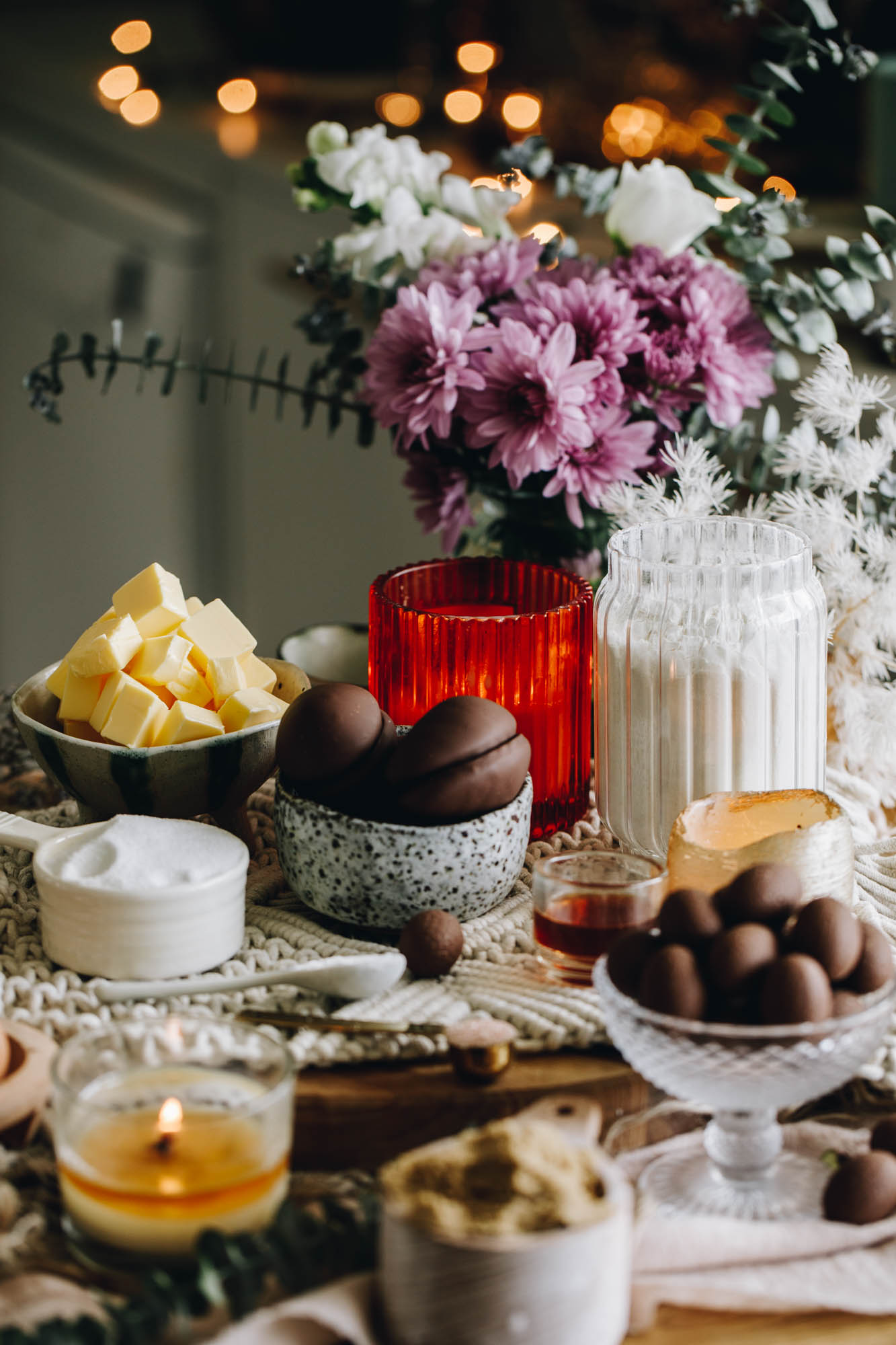 A round wooden board has a cream weaved mat sitting on it. On the board is vintage bowls and cups filled with biscuit ingredients. There is mini easter eggs, marshmallow chocolate eggs, sugar, cubed butter and flour are in view. An orange candle is burning behind the ingredients. A bunch of flowers are also in the background with fairy lights. 