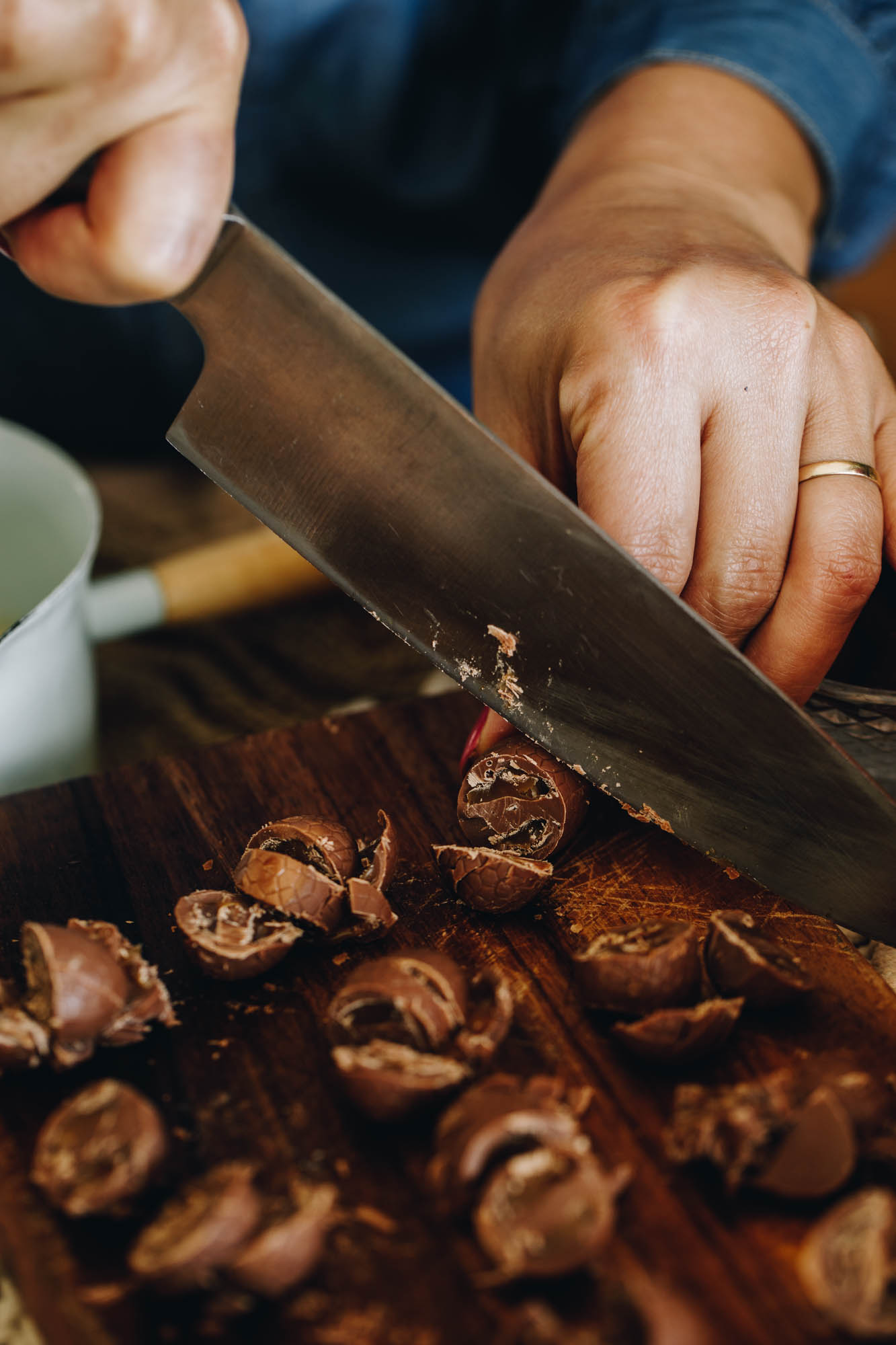 On a wooden chopping board is mini chocolate easter eggs that have been sliced. Naomi is slicing through one with a large silver knife. A small blue pot with a wooden handle is in the background. 