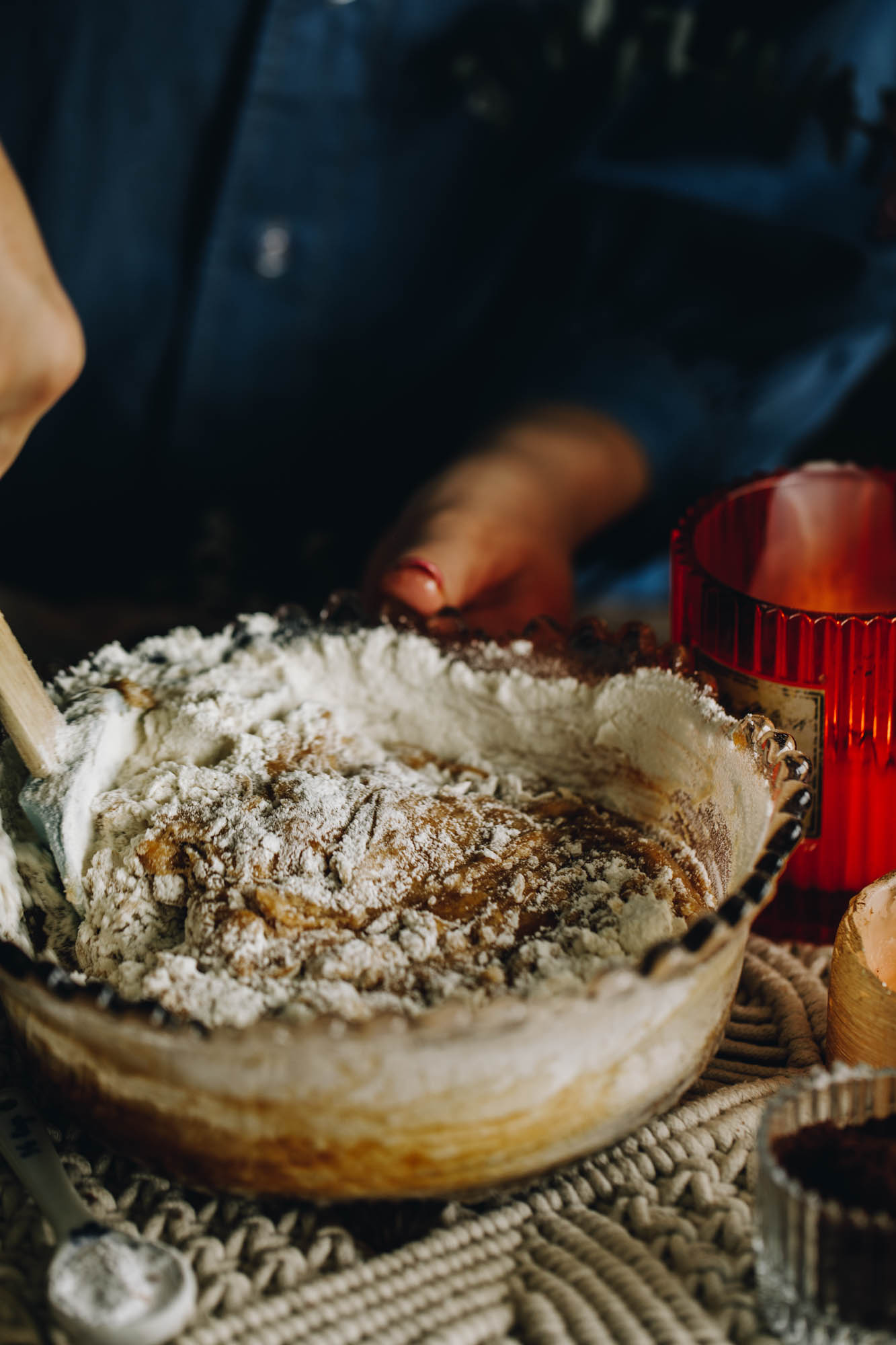 On a cream weaved mat sits a purple vintage bowl with a crinkled trim. In the bowl is cookie dough mixture with flour being stirred through with a spatula. An orange candle burns in the background. 