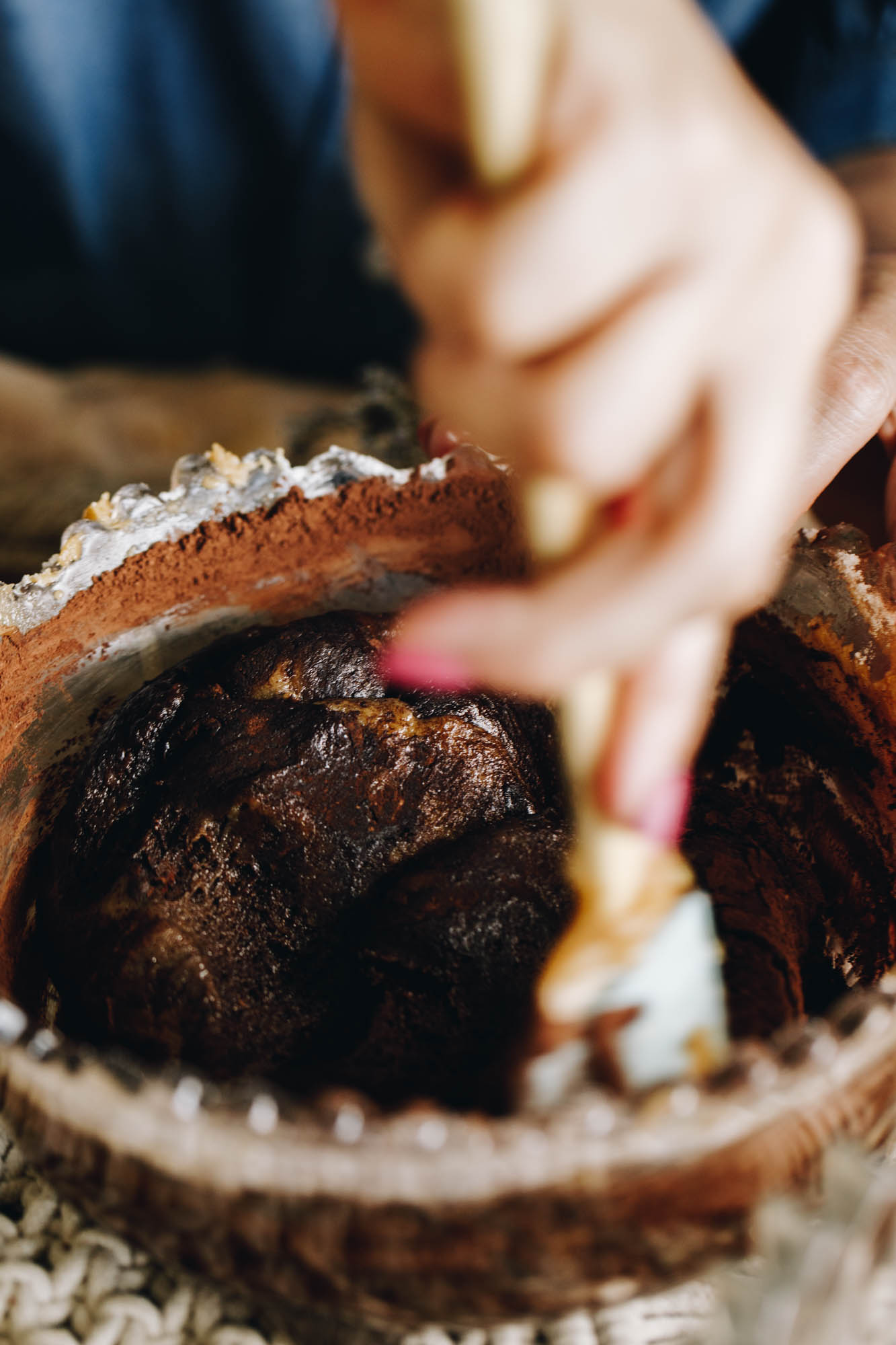 On a cream weaved mat sits a purple vintage bowl with a crinkled trim. In the bowl is a cocoa cookie dough mixture being stirred with a spatula. 