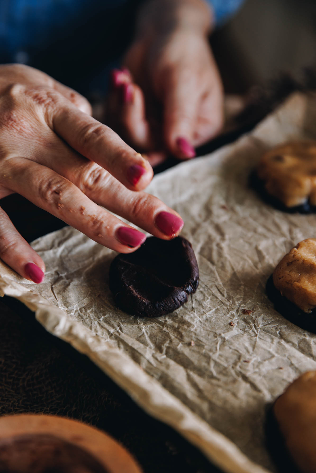 A black tray is lined with brown baking paper. On the paper are cookies and one piece of cocoa dough is being spread out with Naomi's hand. 