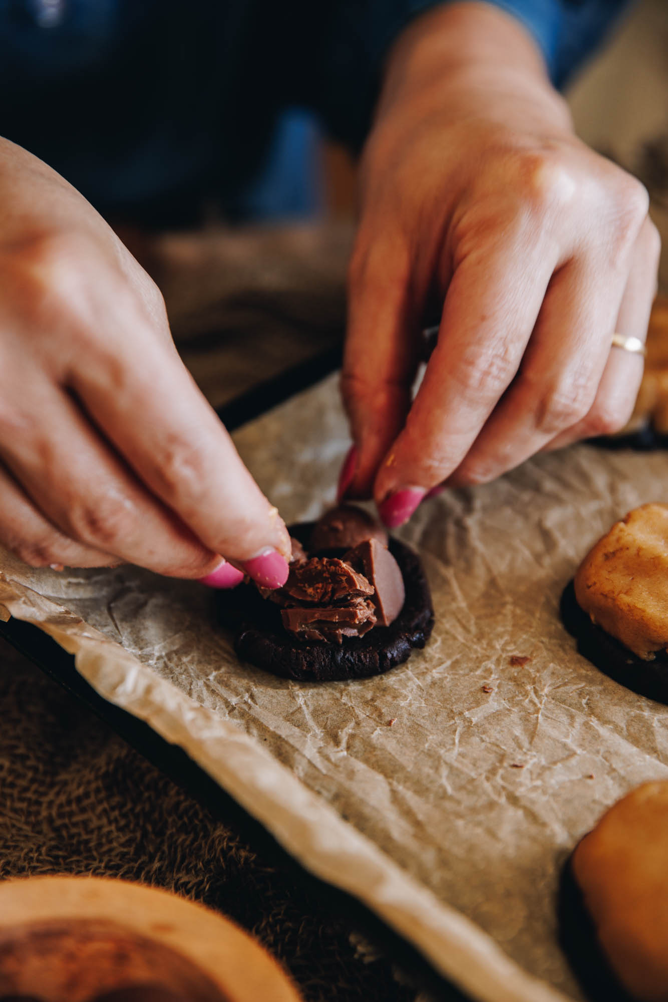 A black tray is lined with brown baking paper. On the paper are cookies and one piece of cocoa dough is having chopped chocolate easter eggs placed in to the centre. 