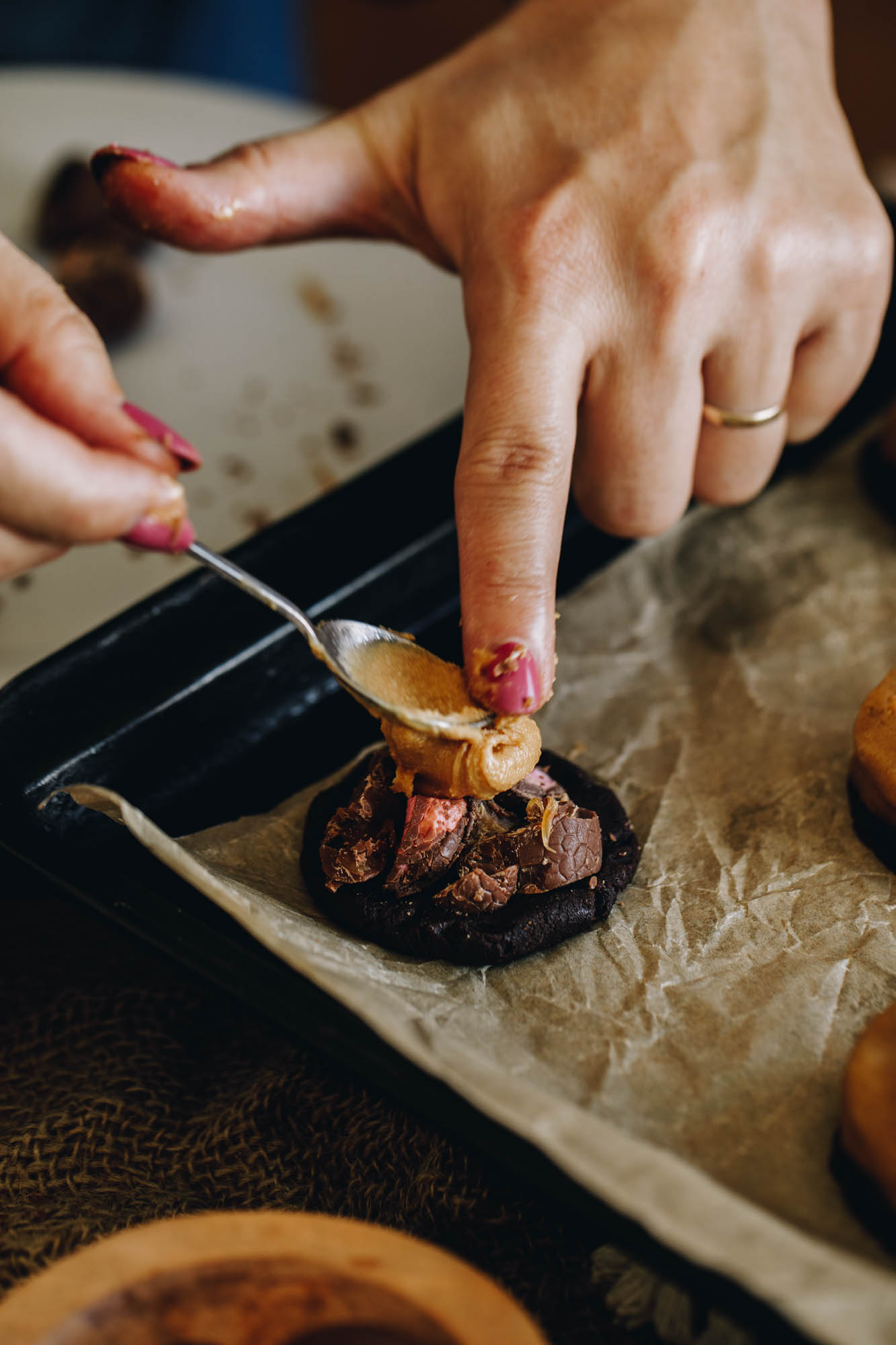 A black tray is lined with brown baking paper. On the paper are cookies and one piece of cocoa dough is having peanut butter added on top of chopped chocolate easter eggs placed in to the centre.