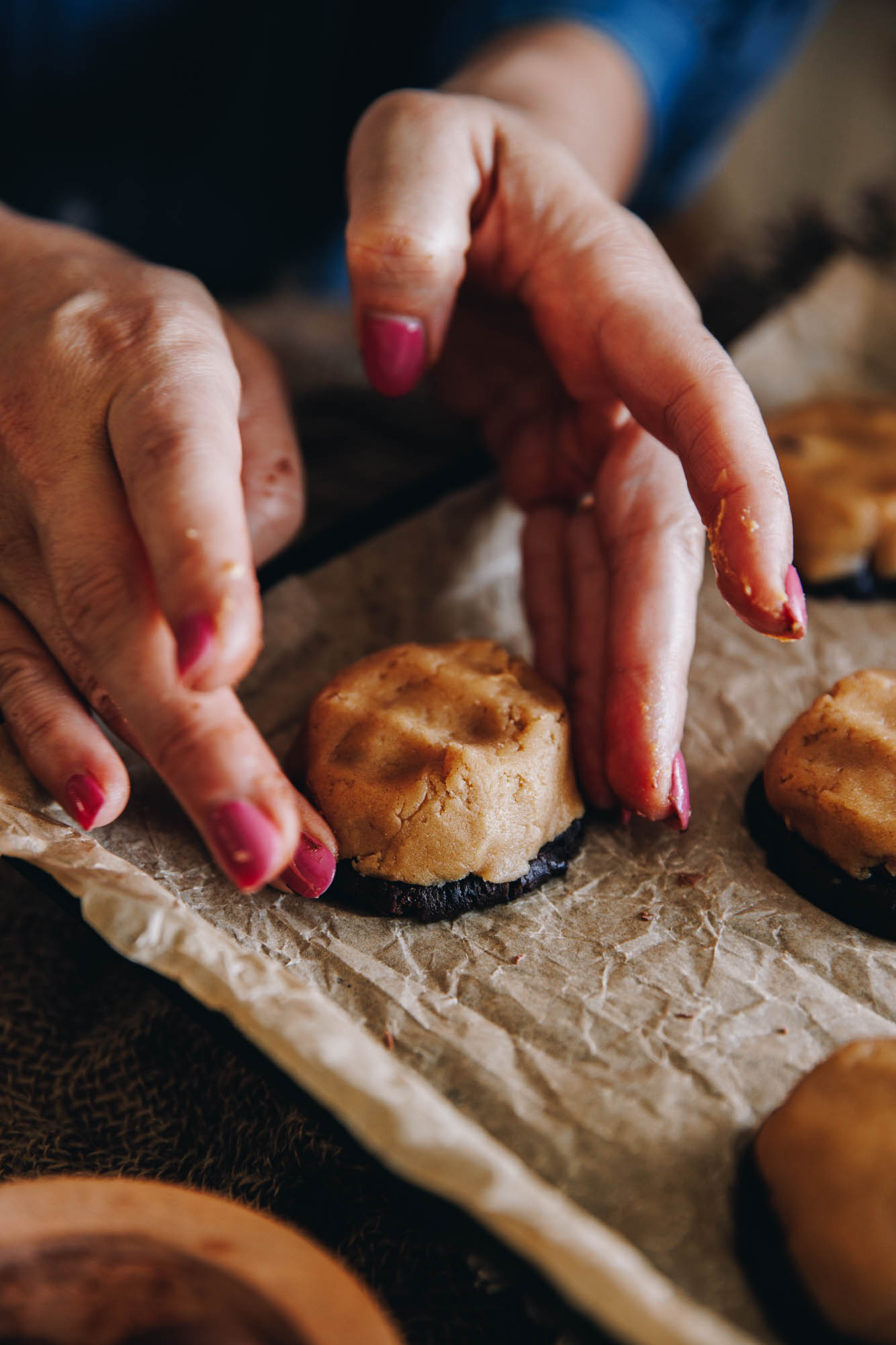 A black tray is lined with brown baking paper. On the paper are cookies and one cookie is having vanilla dough draped over the cocoa biscuit and sealed with Naomi's hand. 