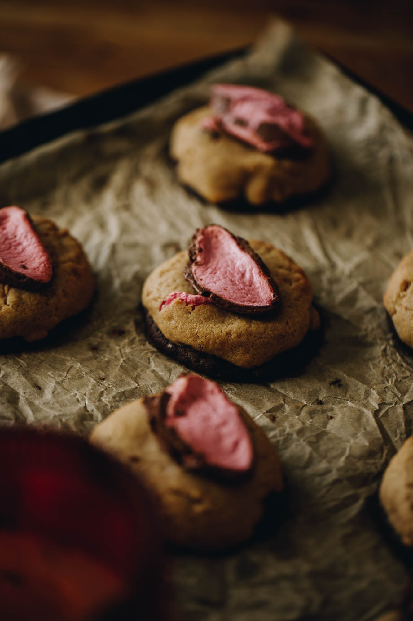 On a black baking tray lined with brown baking paper sits 6 freshly baked chocolate easter egg cookies with pieces of pink chocolate marshmallow baked on the top of each one. The tray is on a wooden table. 