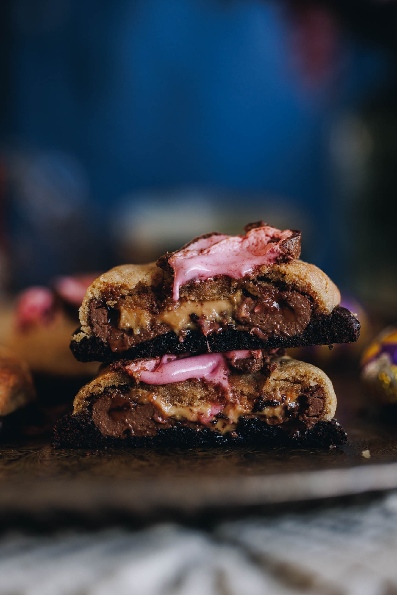 Freshly baked Chocolate easter egg cookies sit on a silver tray. One has been cut in half and stacked on the other half to reveal the gooey chocolate centre. The cookies are two tone, the top layer is vanilla and the bottom layer is chocolate. On top of the cookies is melted pink marshmallow. A weaved mat is under the tray.