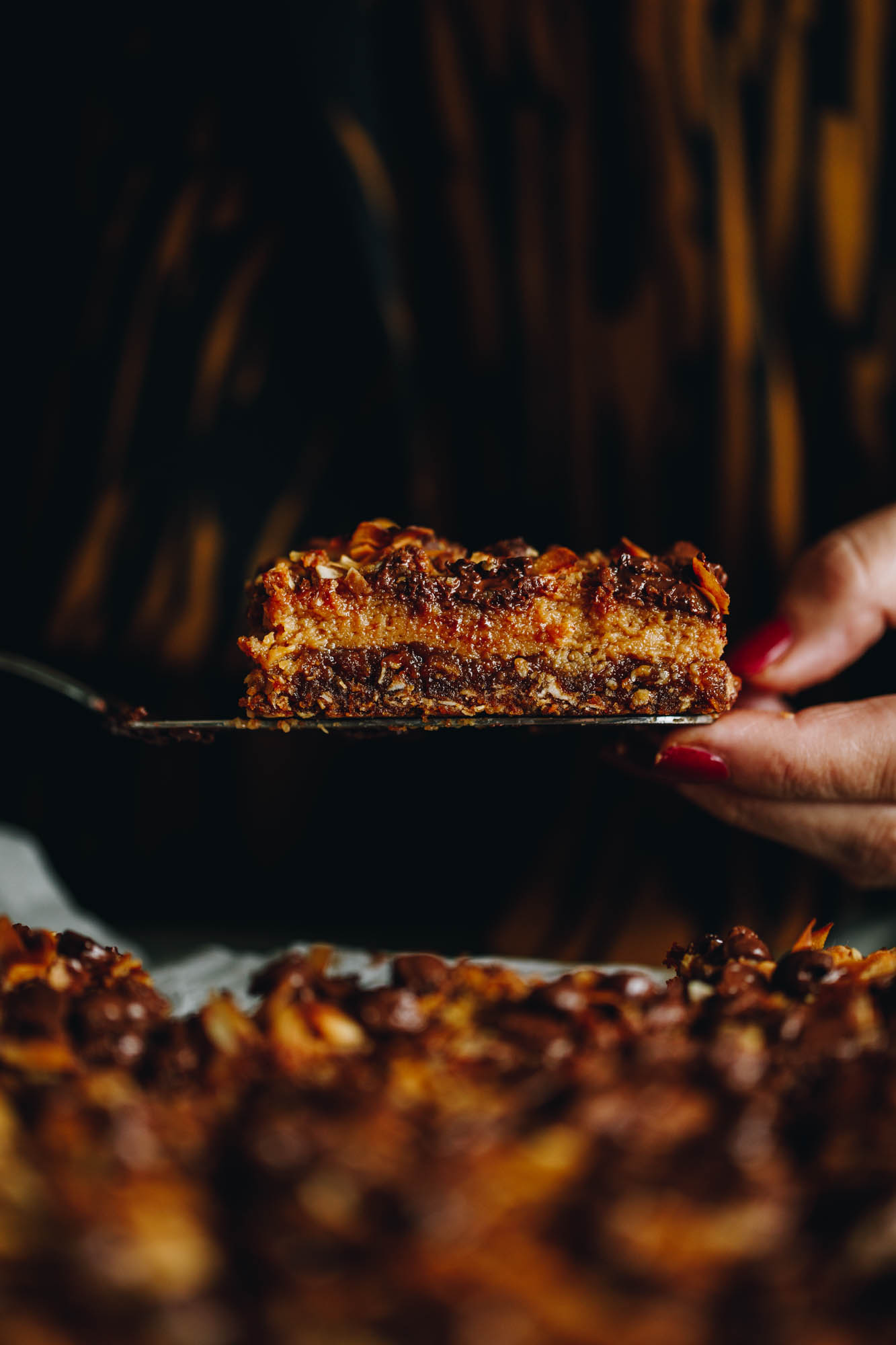 A piece of Best Oaty Caramel Slice is being held up in Naomi's hand on one side and a serve knife on the other. In the forefront is blurred oats caramel slice. Naomi is wearing a striped black and brown top to mimic the same colours as the slice. 