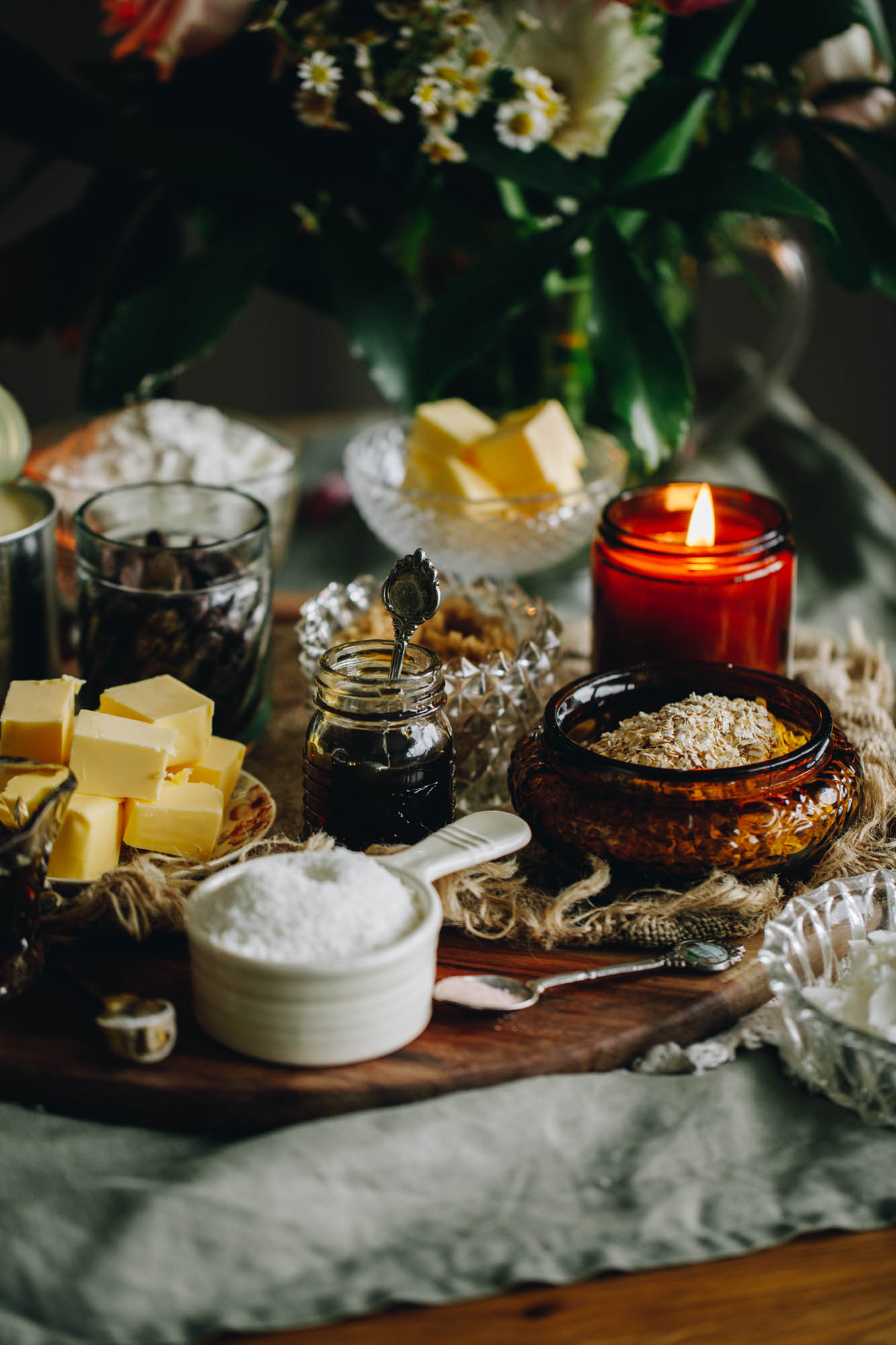 A round wooden board sits on a green cloth, on a wooden table. On the board is vintage cups and vases filled with ingredients. There is sugar, oats, butter, vanilla, golden syrup and flour are in view. An amber candle is burning on it too. In the background is a glass vase with flowers in it.