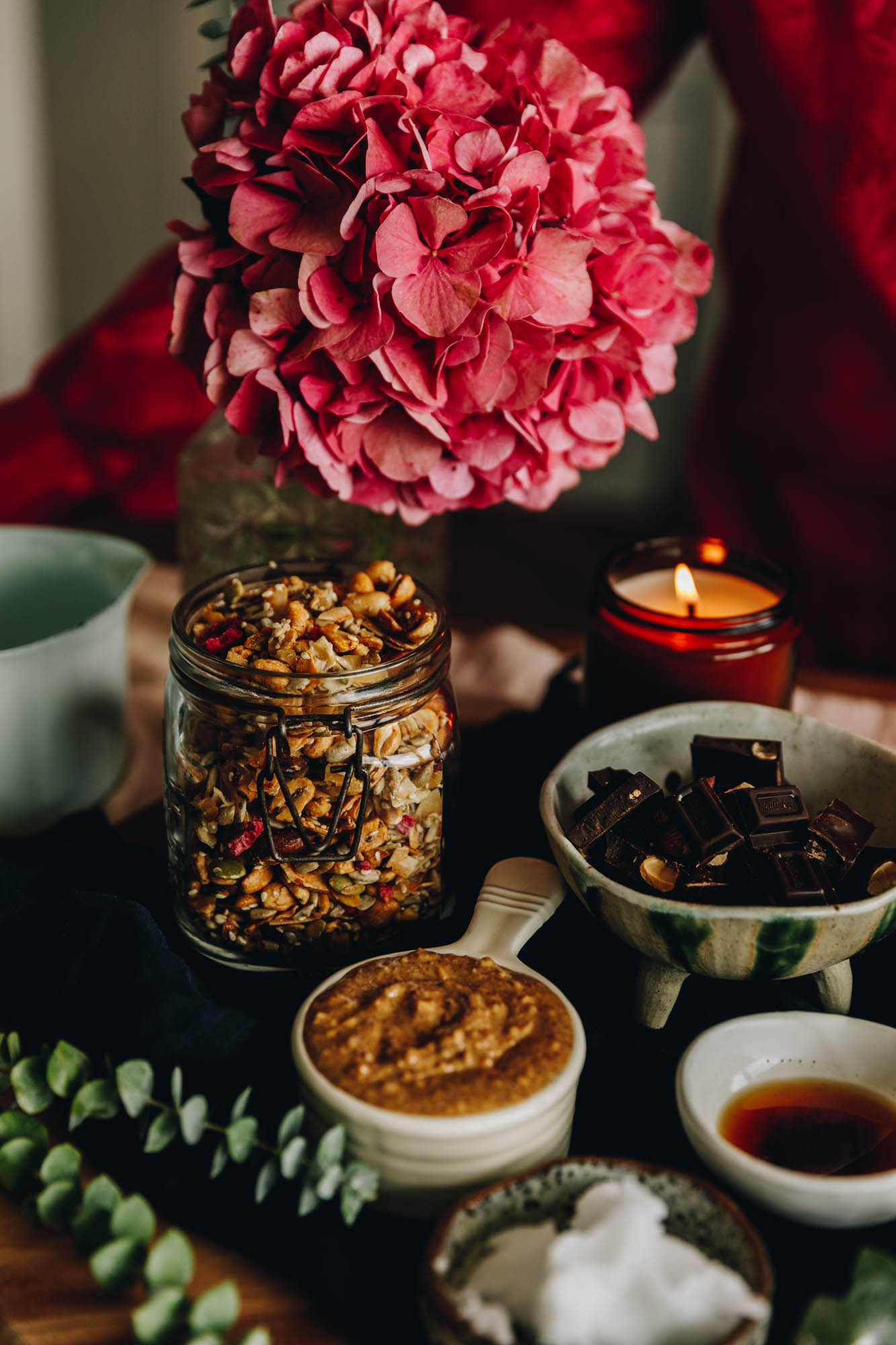 Ingredients are in different vintage bowls and are sitting on a wooden board on top of wooden table. There is a large pink hydrangea n the background and an amber candle burning. 