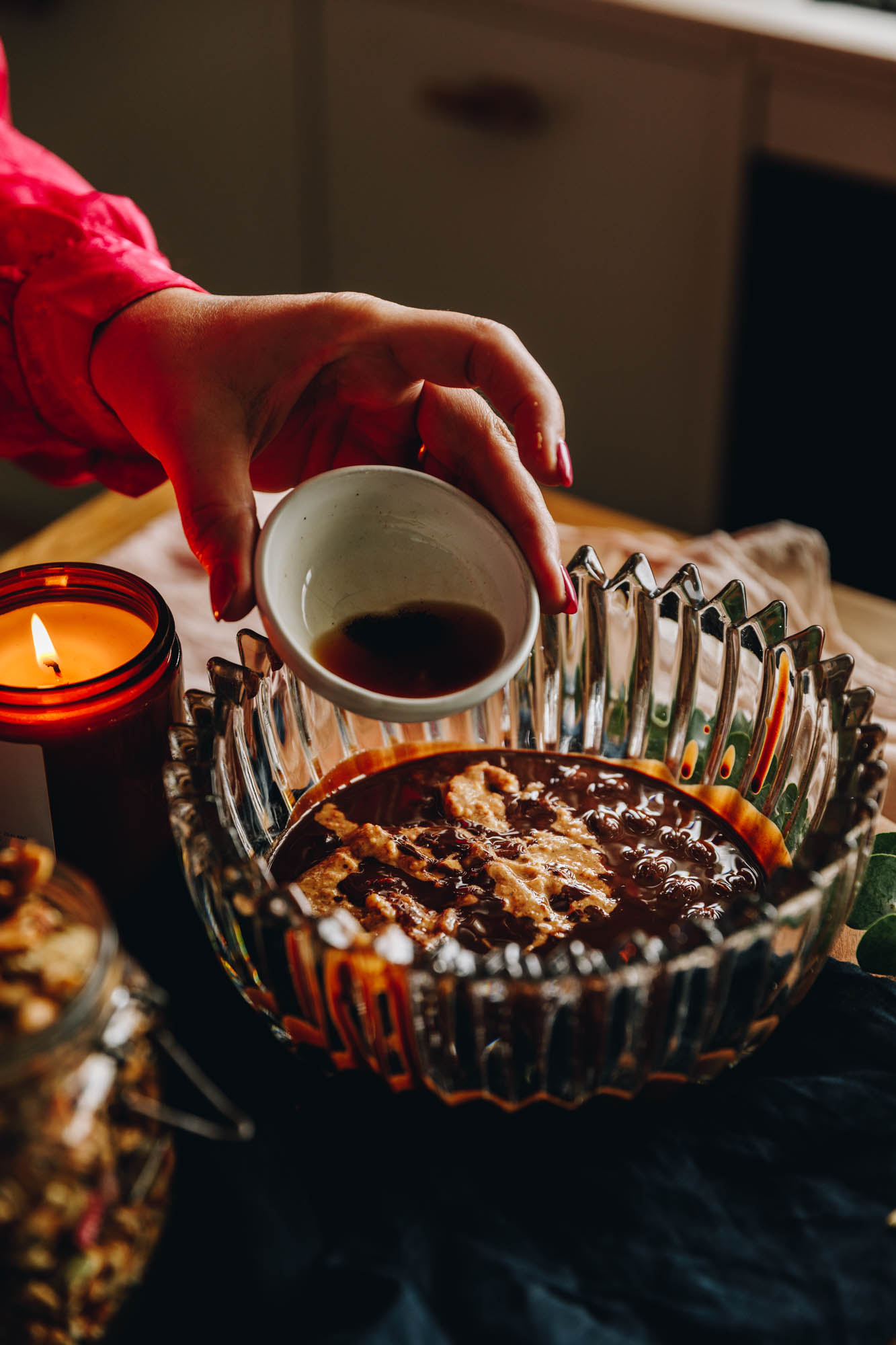 On a wooden table, a vintage bowl sits on it with an amber candle burning in the background. In the bowl with melted chocolate and almond butter. Vanilla is being added in to it. 