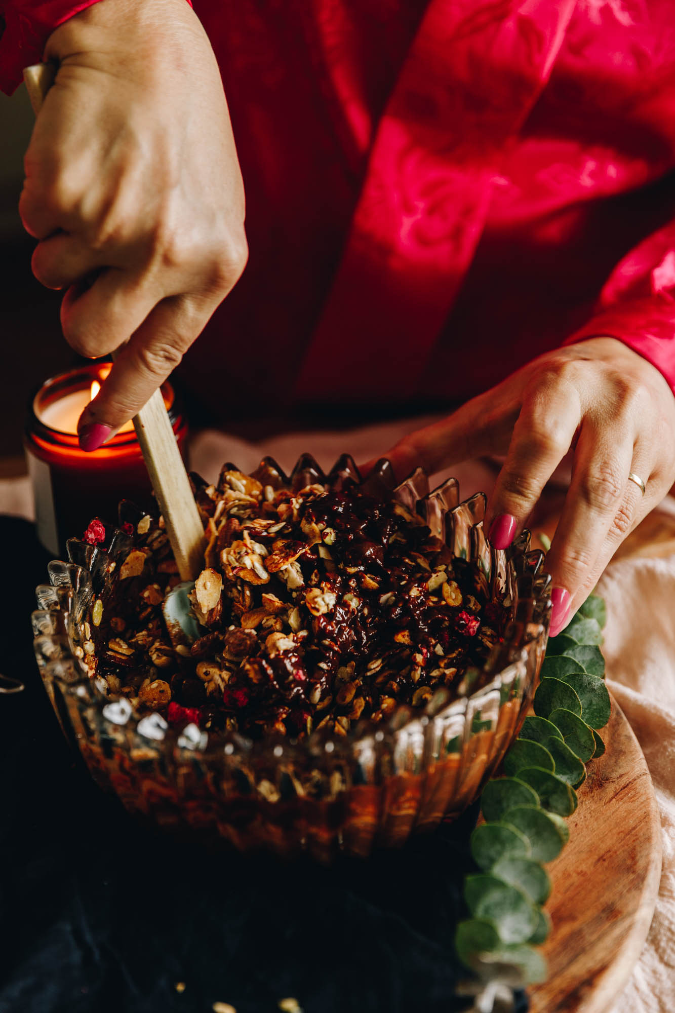 On a wooden table, a vintage bowl sits on it with an amber candle burning in the background. In the bowl all of the chocolate nut and seed slice mixture, it is being stirred with a spatula. 