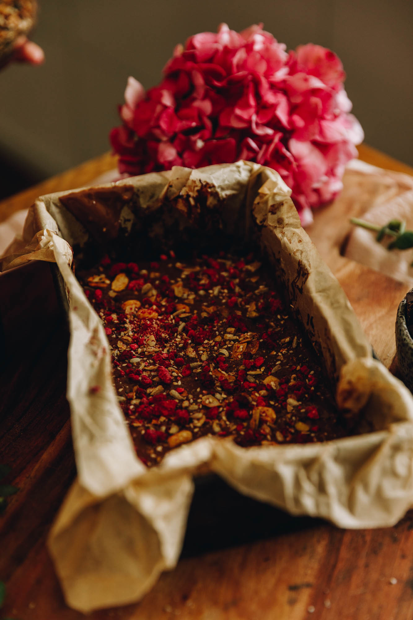 A tin lined with brown paper and the chocolate and seed mixture has been pressed in.It has been topped with more chocolate and seeds and freeze dried fruit sprinkles. It sits on a wooden table.