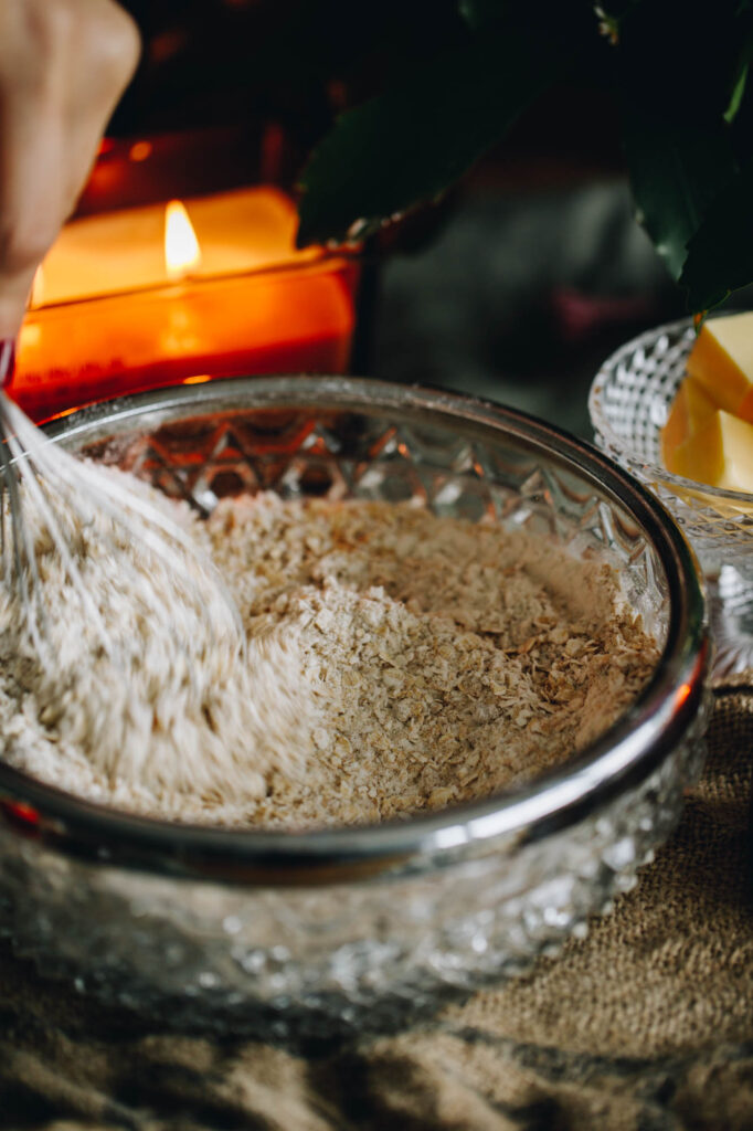 In a glass bow with a silver trim is oats and flour being mixed together with a whisk. A candle is burning in the background. 