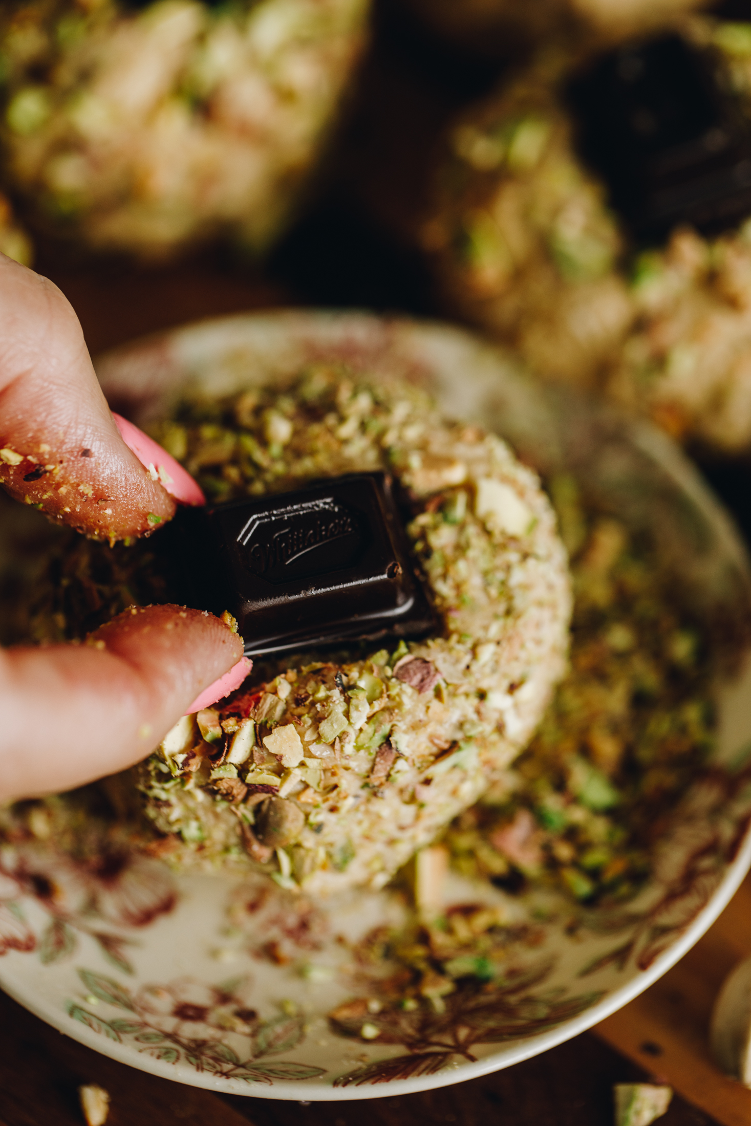 Close shot of a piece of dark chocolate being placed in the centre of a pistachio and chocolate cookie. It is on a small vintage plate. 