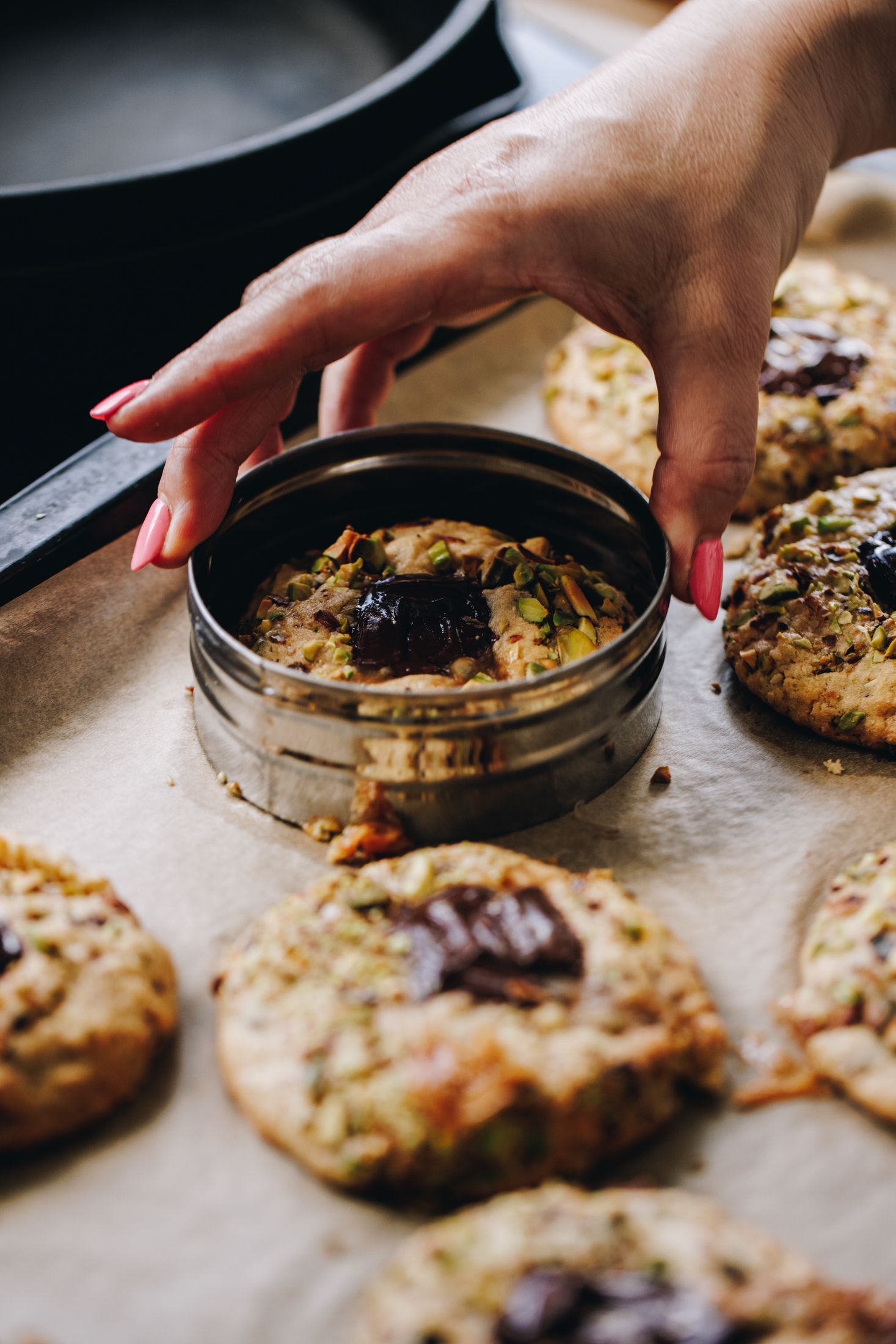 Freshly baked pistachio and chocolate cookies are on a tray with brown baking paper. A hand is using a large cookie cutter to shape a cookie. 
